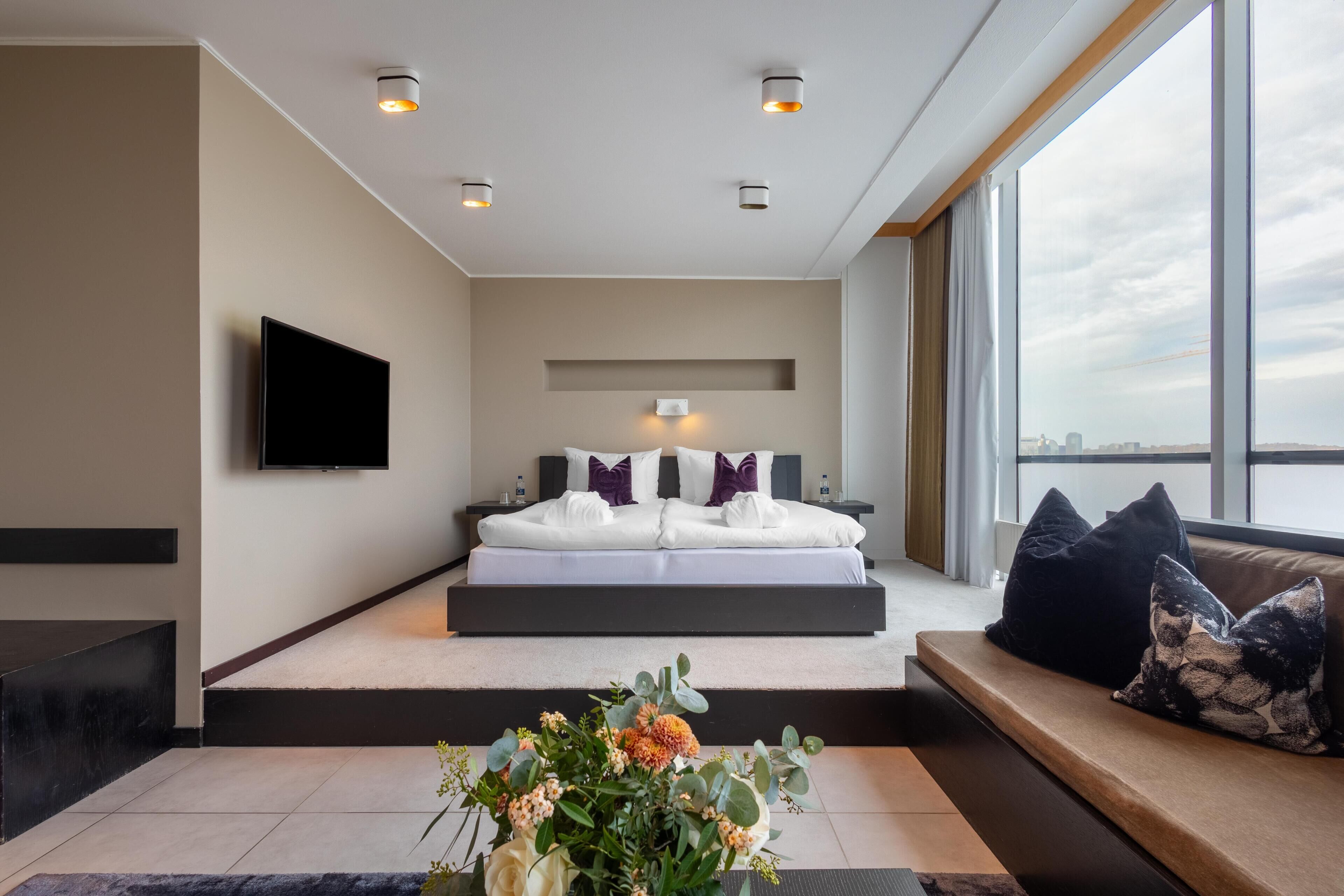 A modern bedroom with a large bed, a flat-screen TV, a view of the city, and a small table with flowers.