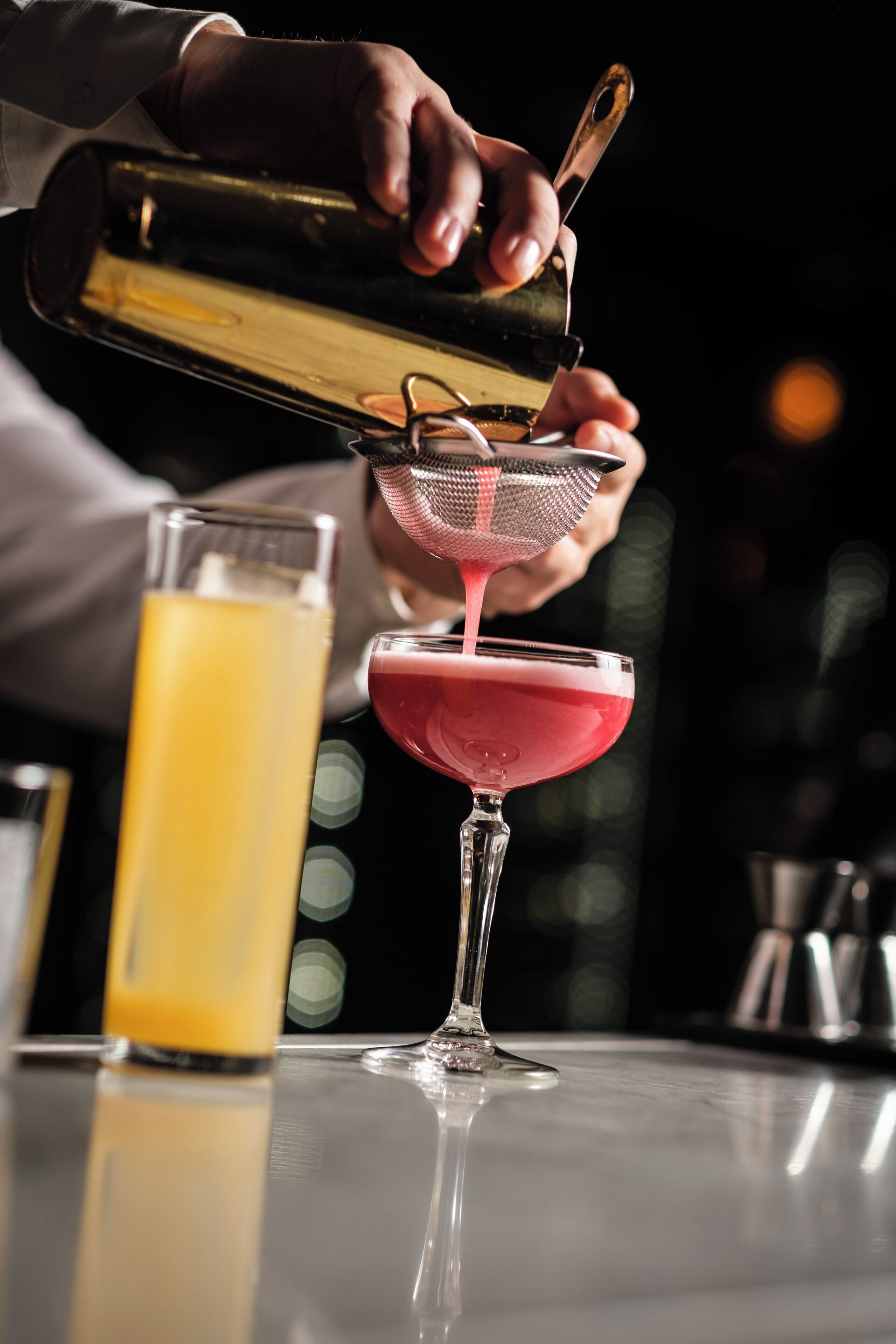 A bartender pouring a pink liquid into a martini glass using a strainer.