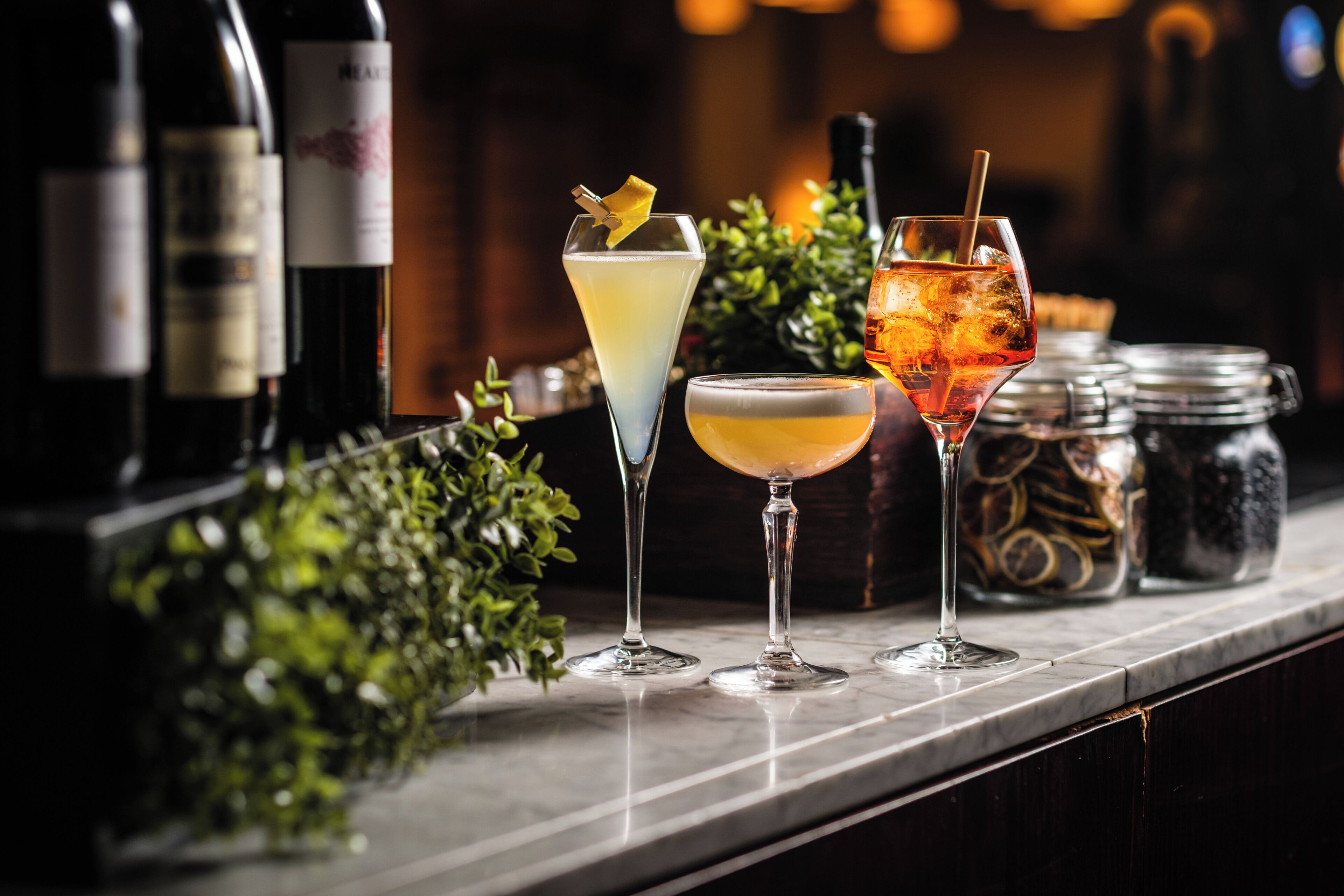Three cocktails on a bar counter with bottles and jars in the background.