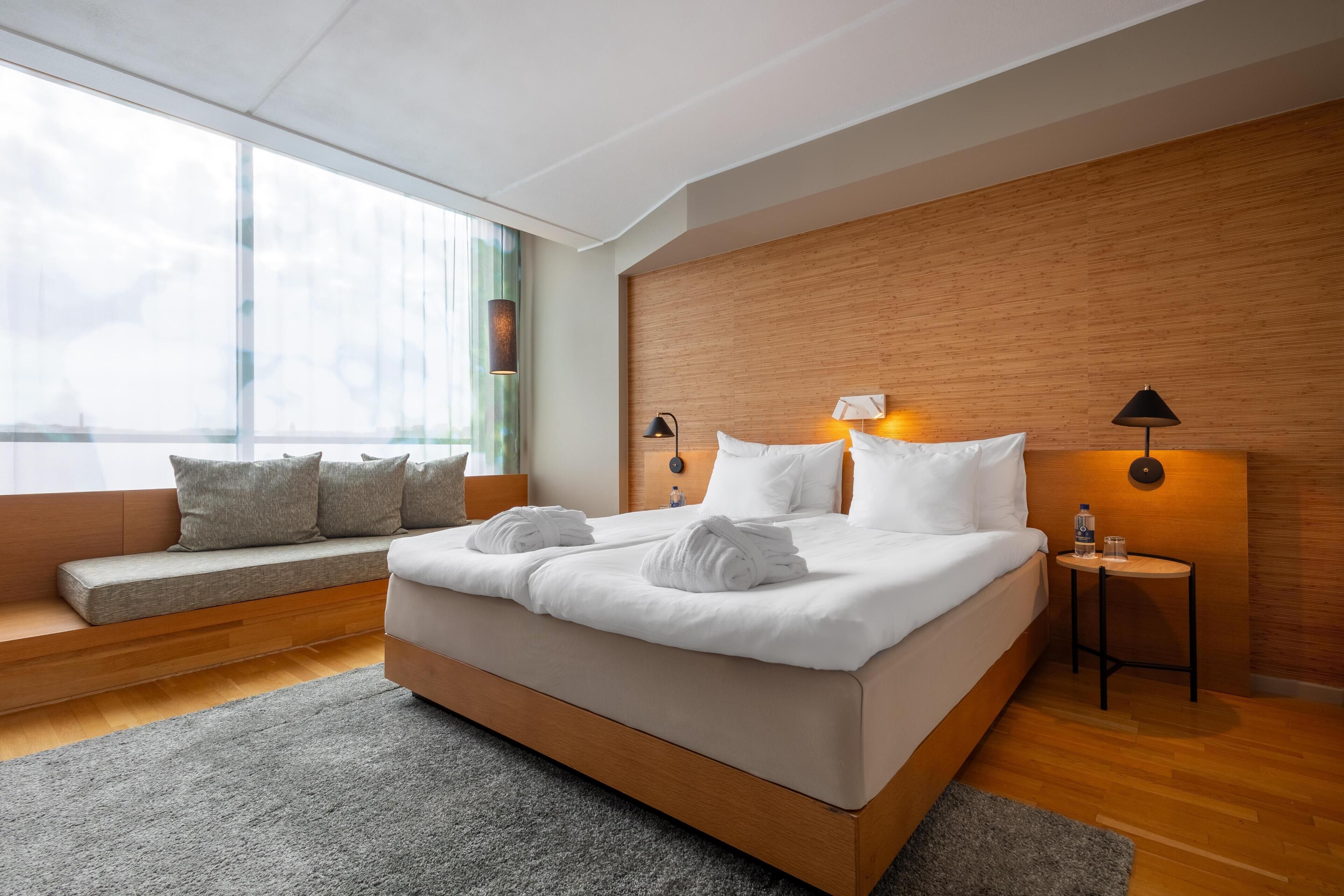 A modern hotel room with a large bed, a seating area with cushions, a wooden headboard, and a view of the outdoors.