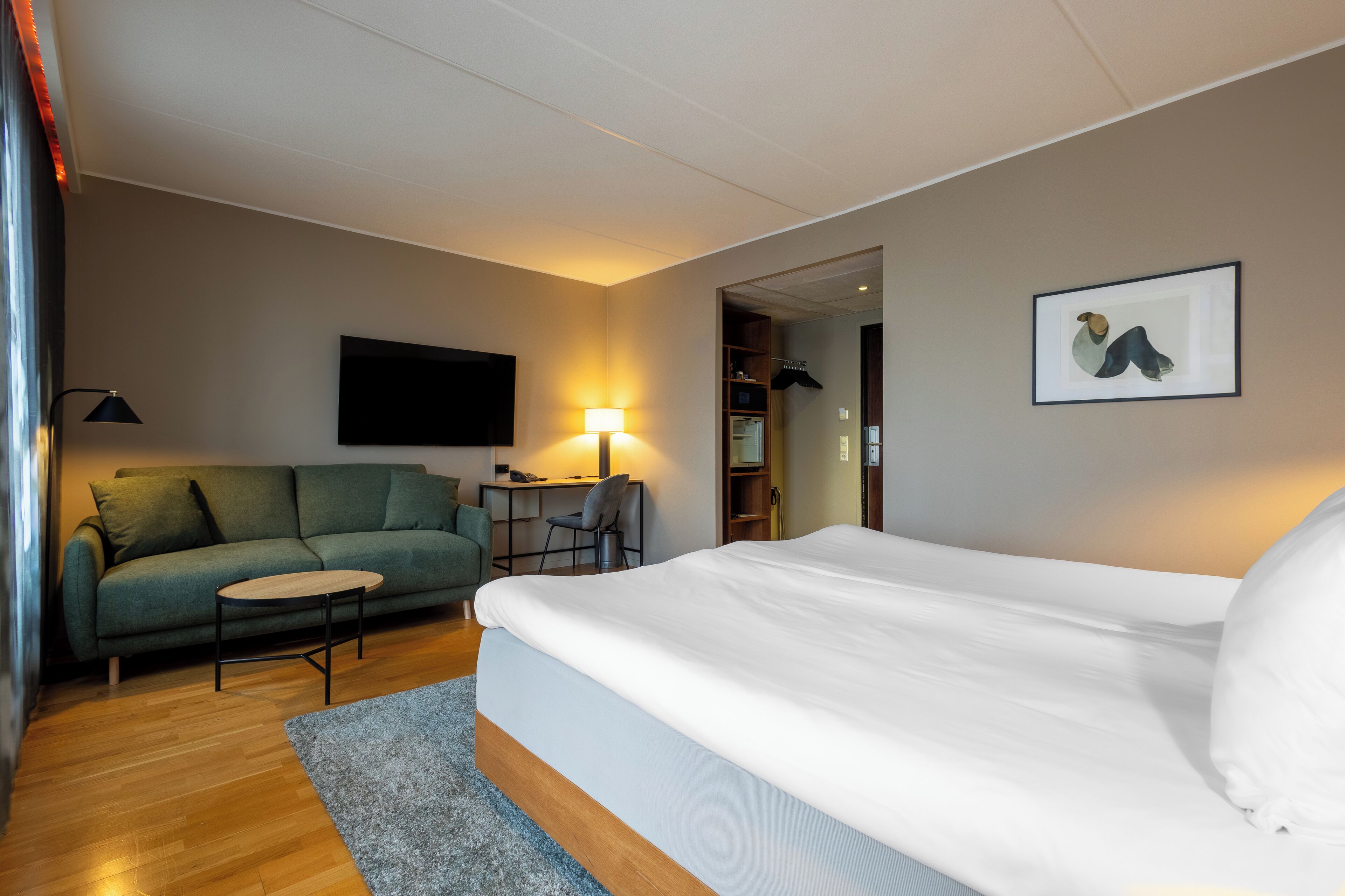 A hotel room with a bed, a green sofa, a small table, a lamp, and a framed picture on the wall.