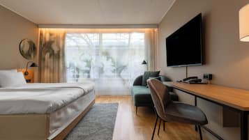 Quarto de hotel moderno com cama, escrivaninha, cadeira, televisão e janela grande com cortinas.