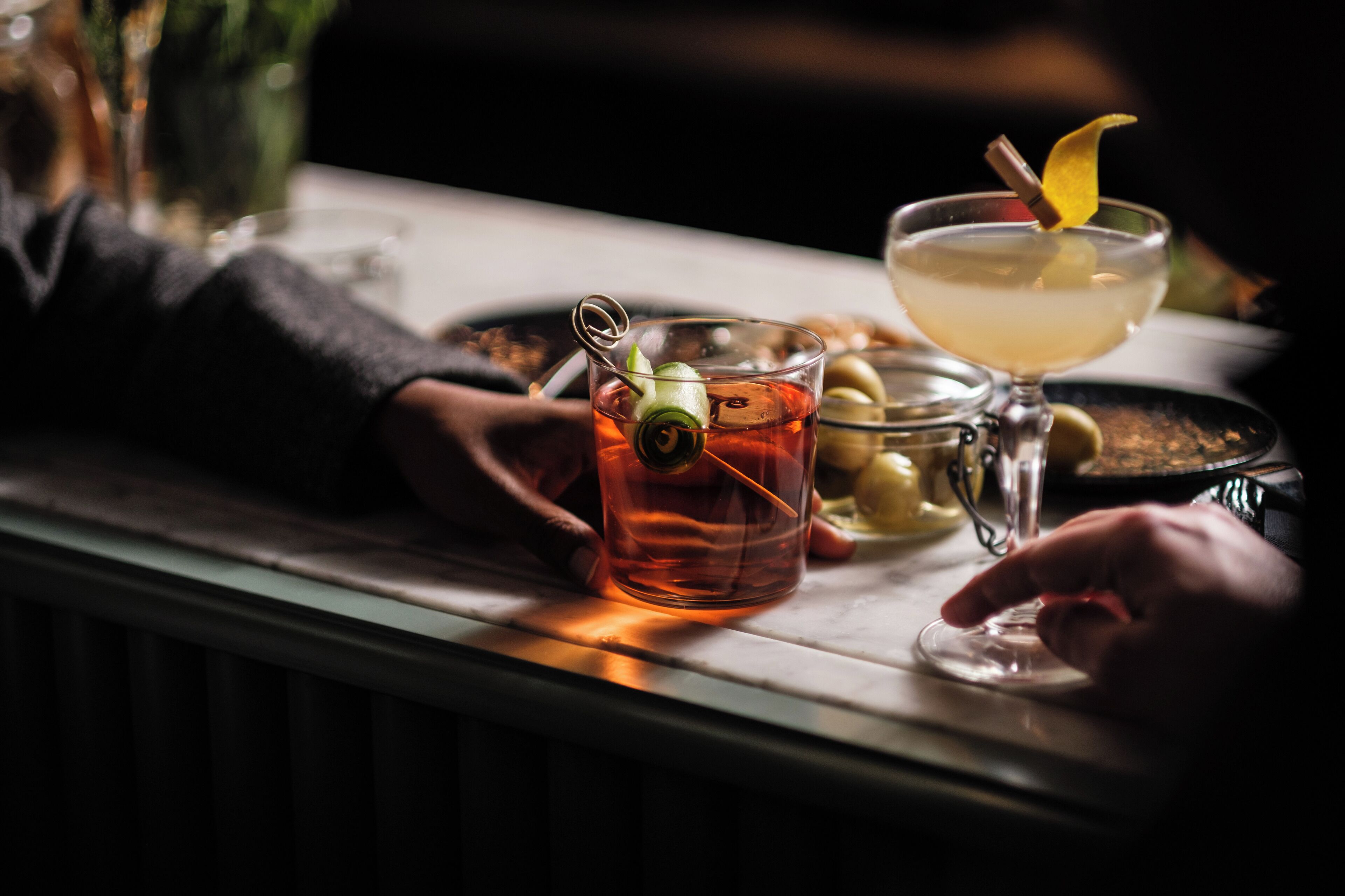 A bar setting with a glass of amber liquid and a cocktail with a garnish.