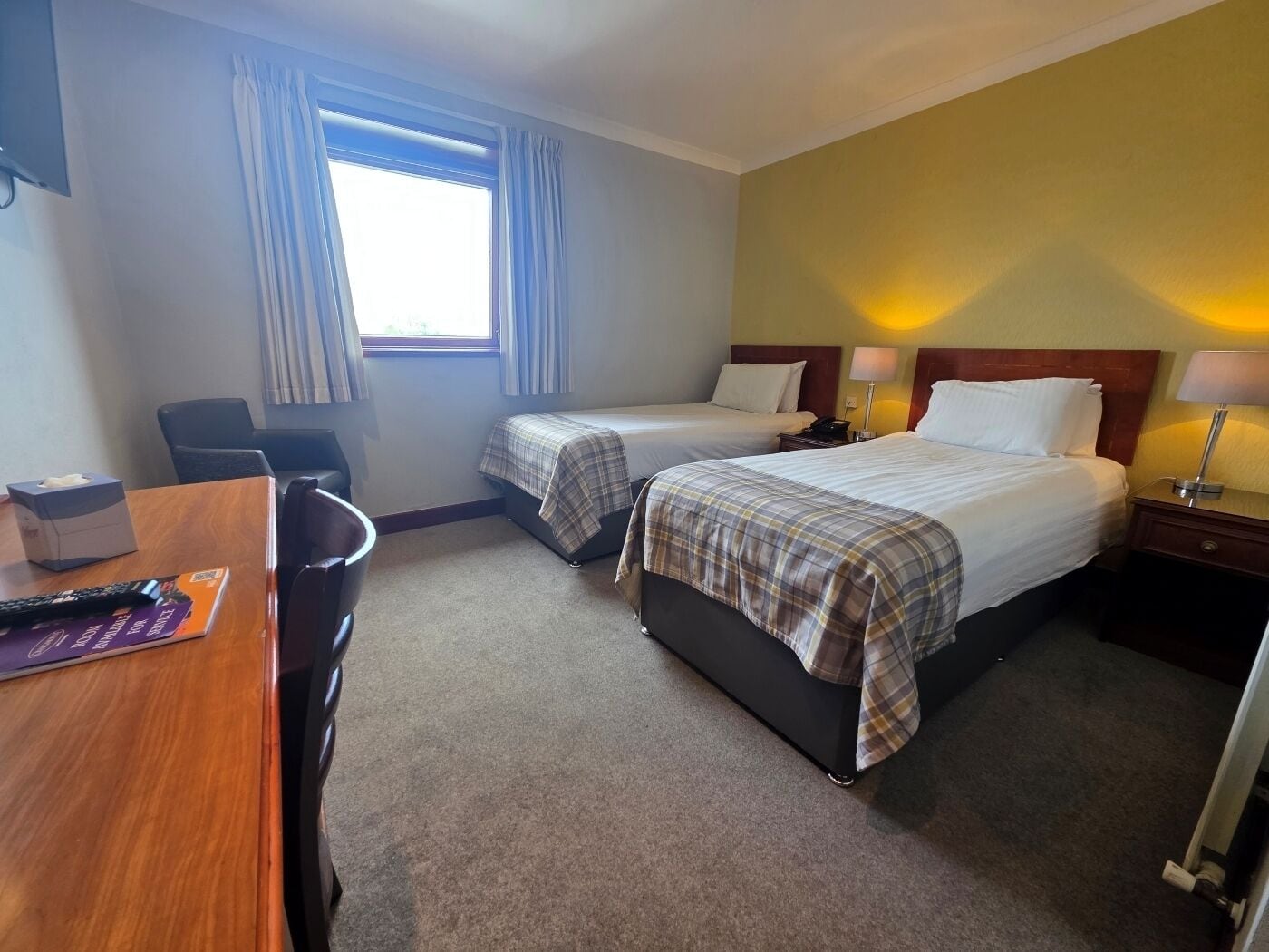 Une chambre d’hôtel avec deux lits, un bureau, une chaise, une fenêtre avec des rideaux et une lampe de chevet.