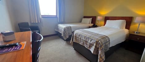 A hotel room with two beds, a desk, a chair, a window with curtains, and a bedside lamp.