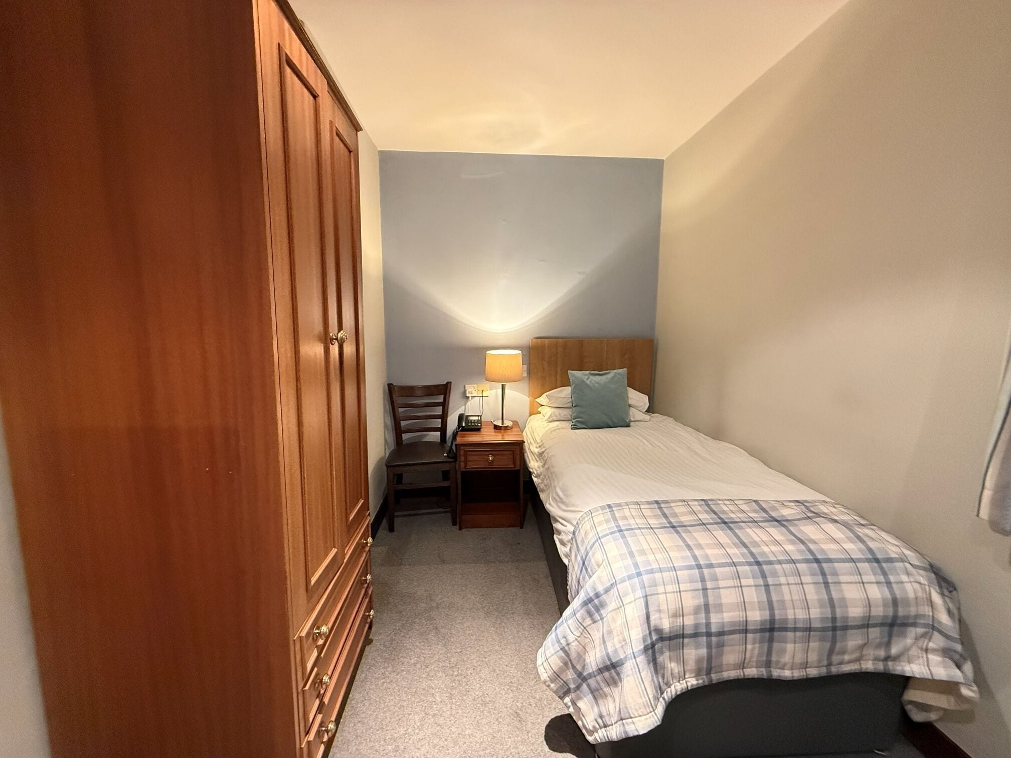 A small, single-bed room with a wooden wardrobe, a bedside table with a lamp, and a chair.
