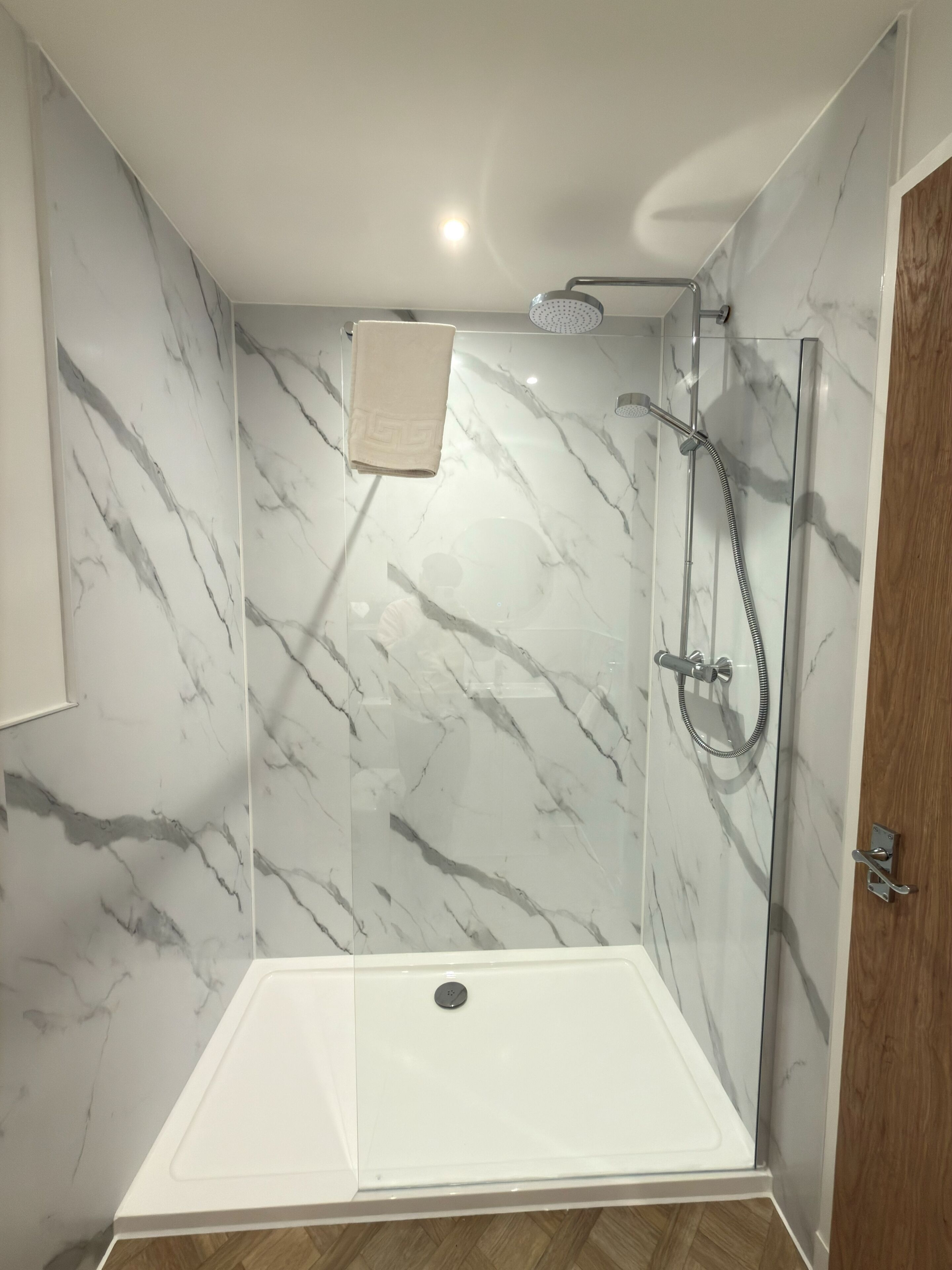 A walk-in shower with a glass door, a towel hanging on the wall, and a wooden floor.
