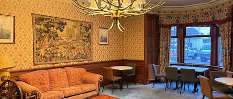 Une pièce avec un papier peint à motifs, un grand tableau, un lustre, un canapé, une table basse et un coin repas avec des chaises et des tables.