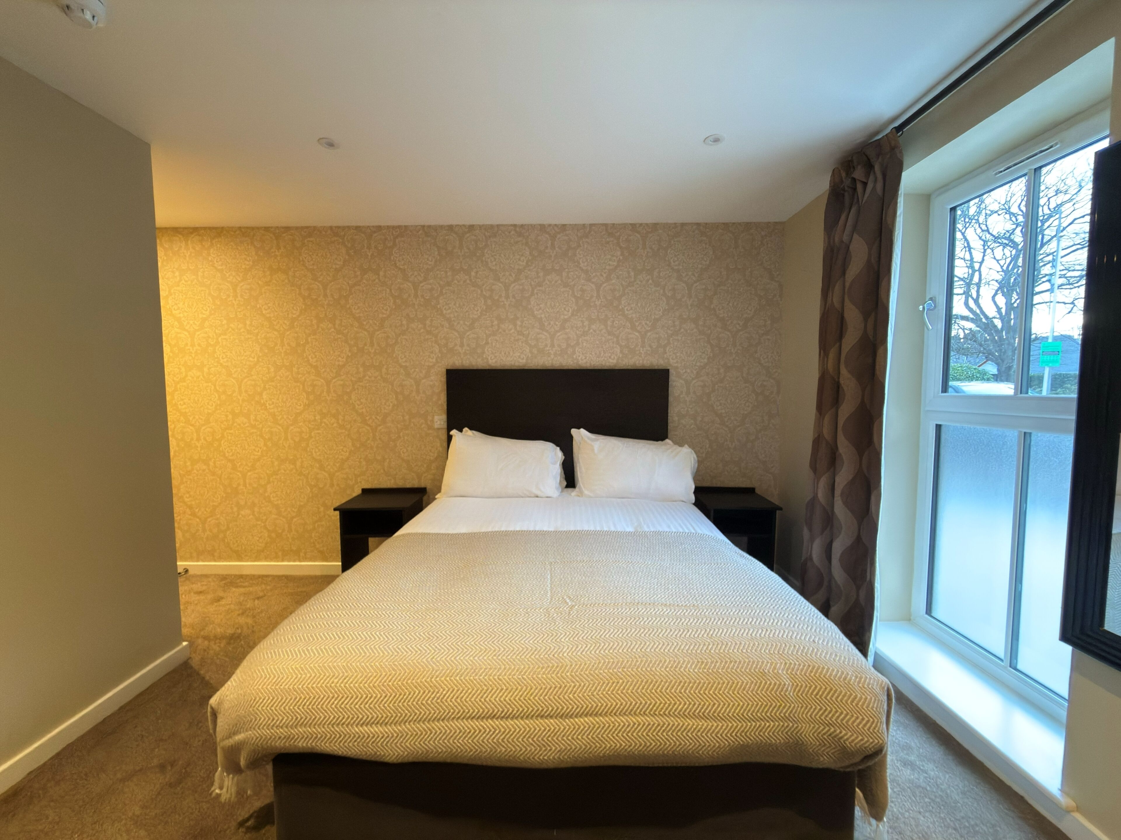 A hotel room with a large bed, two bedside tables, a window with curtains, and patterned wallpaper.