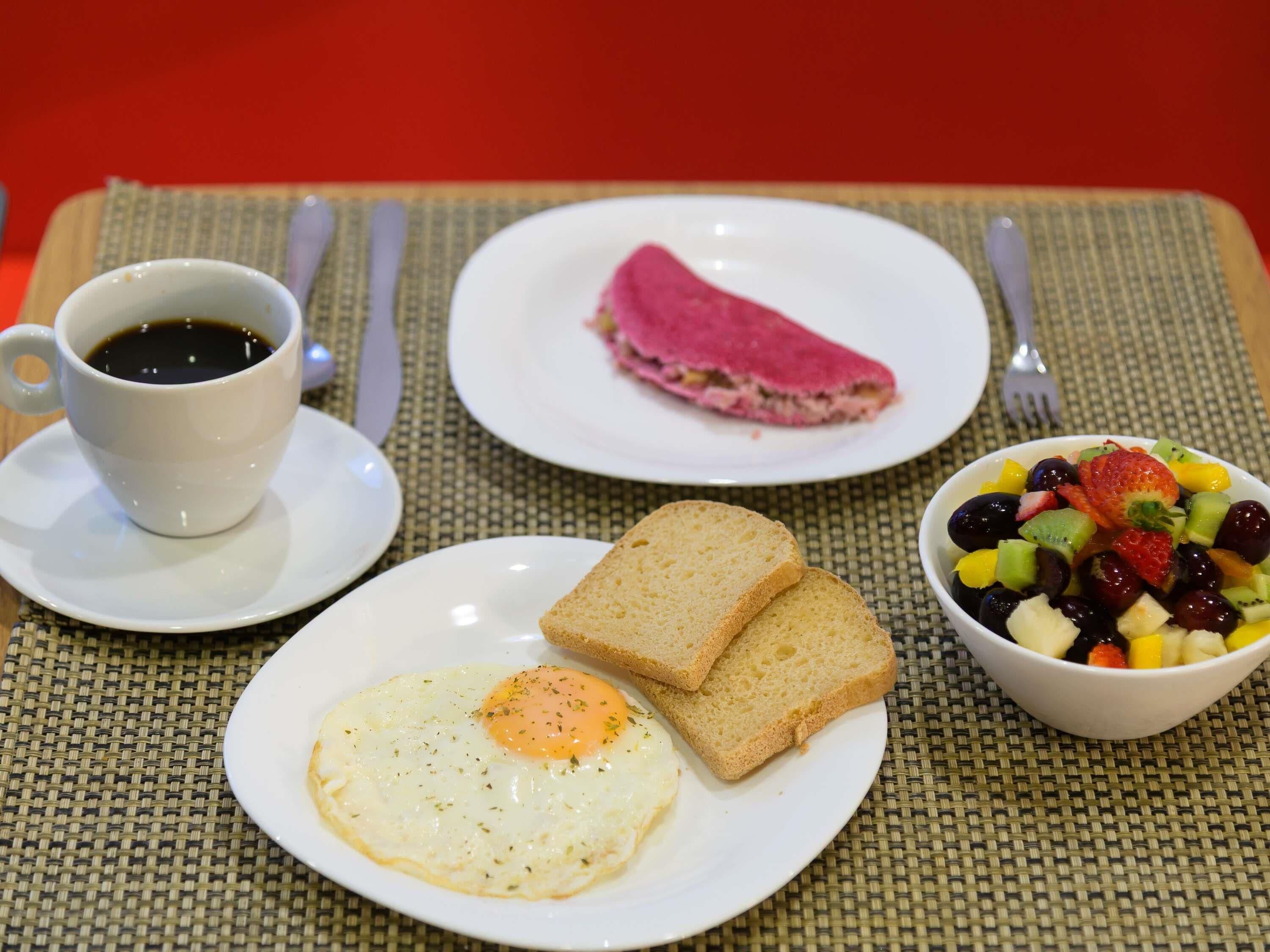 Un desayuno con una taza de café, un plato de tostadas y un huevo, una porción de pastel rosa y un tazón de ensalada de frutas.