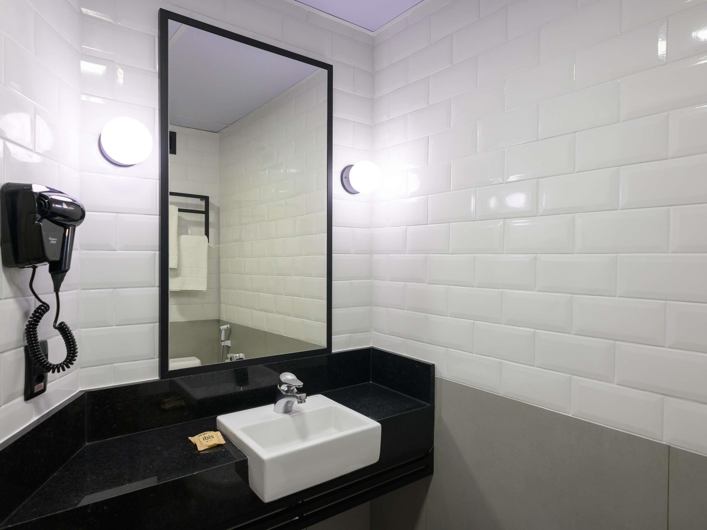 Un baño con paredes de azulejos blancos, encimera negra, lavabo blanco, espejo y secador de pelo.