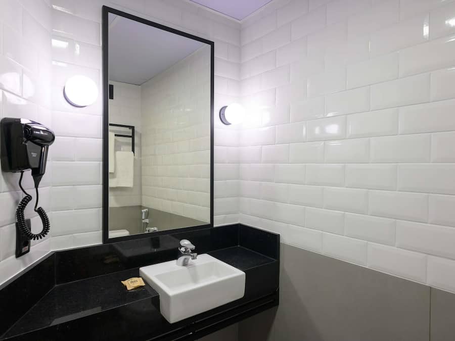 Un baño con paredes de azulejos blancos, encimera negra, lavabo blanco, espejo y secador de pelo.