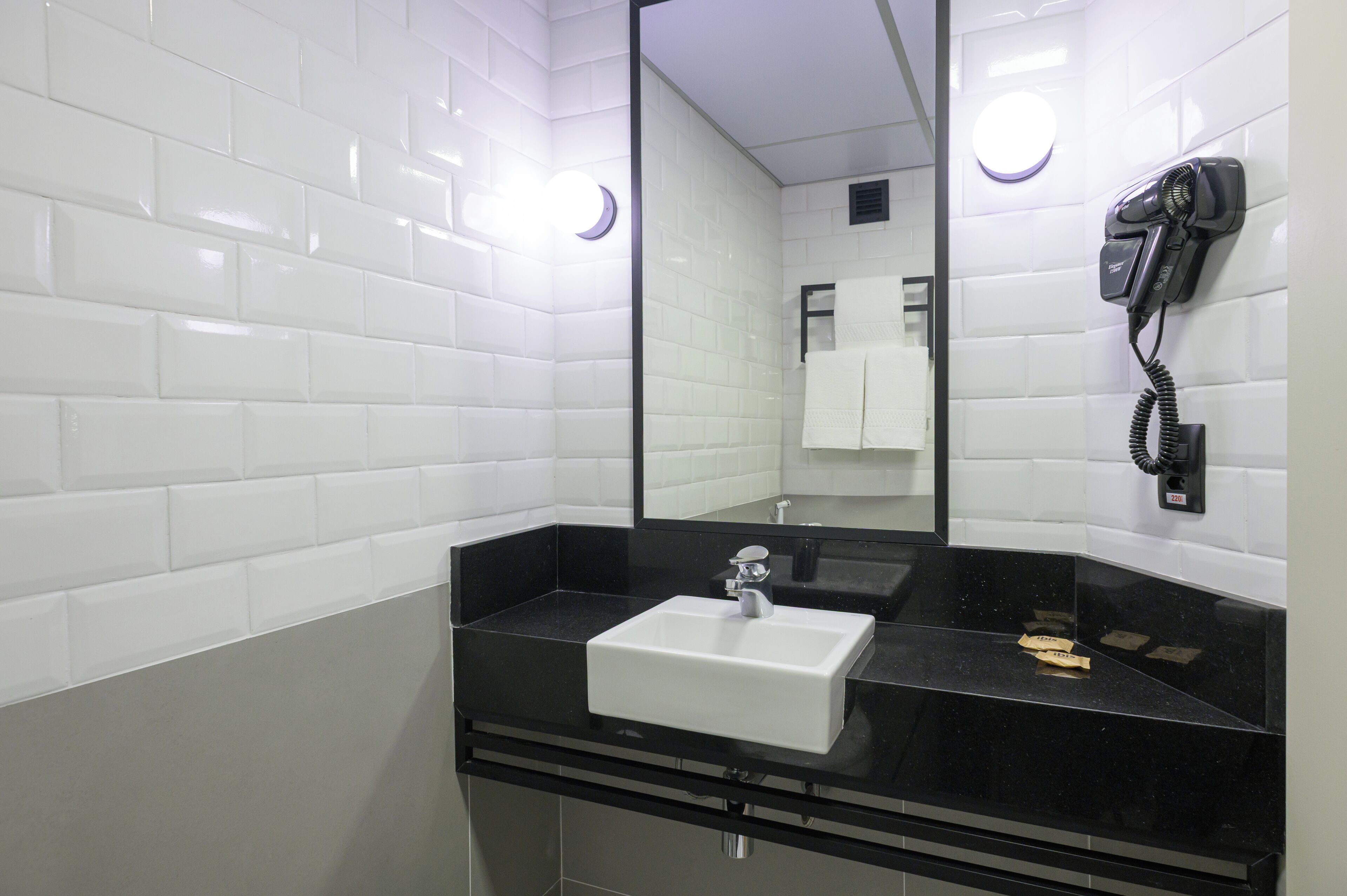 Un baño moderno con azulejos blancos tipo metro, encimera negra, lavamanos blanco y un secador de pelo montado en la pared.
