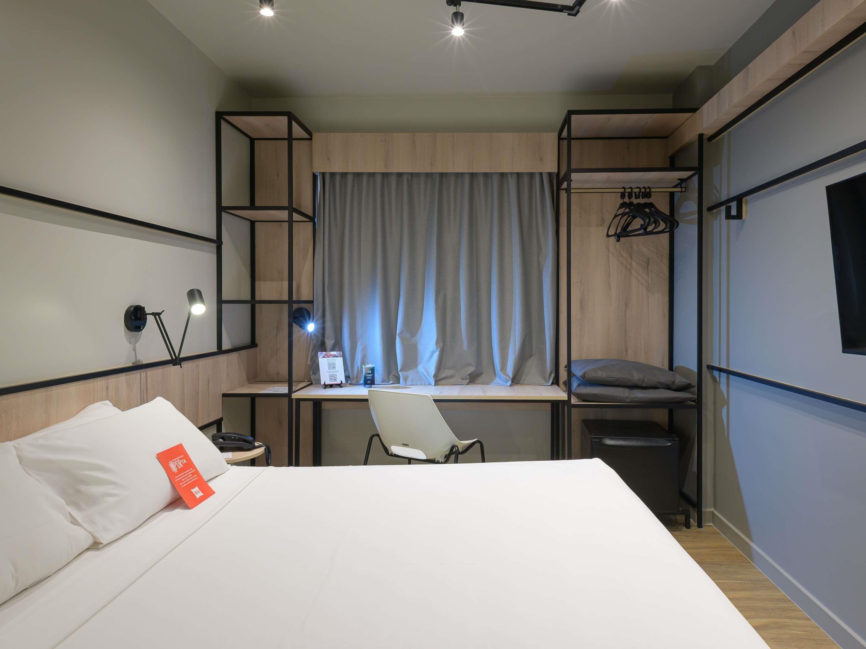 Une chambre d’hôtel moderne équipée d’un lit, d’un bureau, d’une chaise et d’une armoire où sont suspendus des vêtements.
