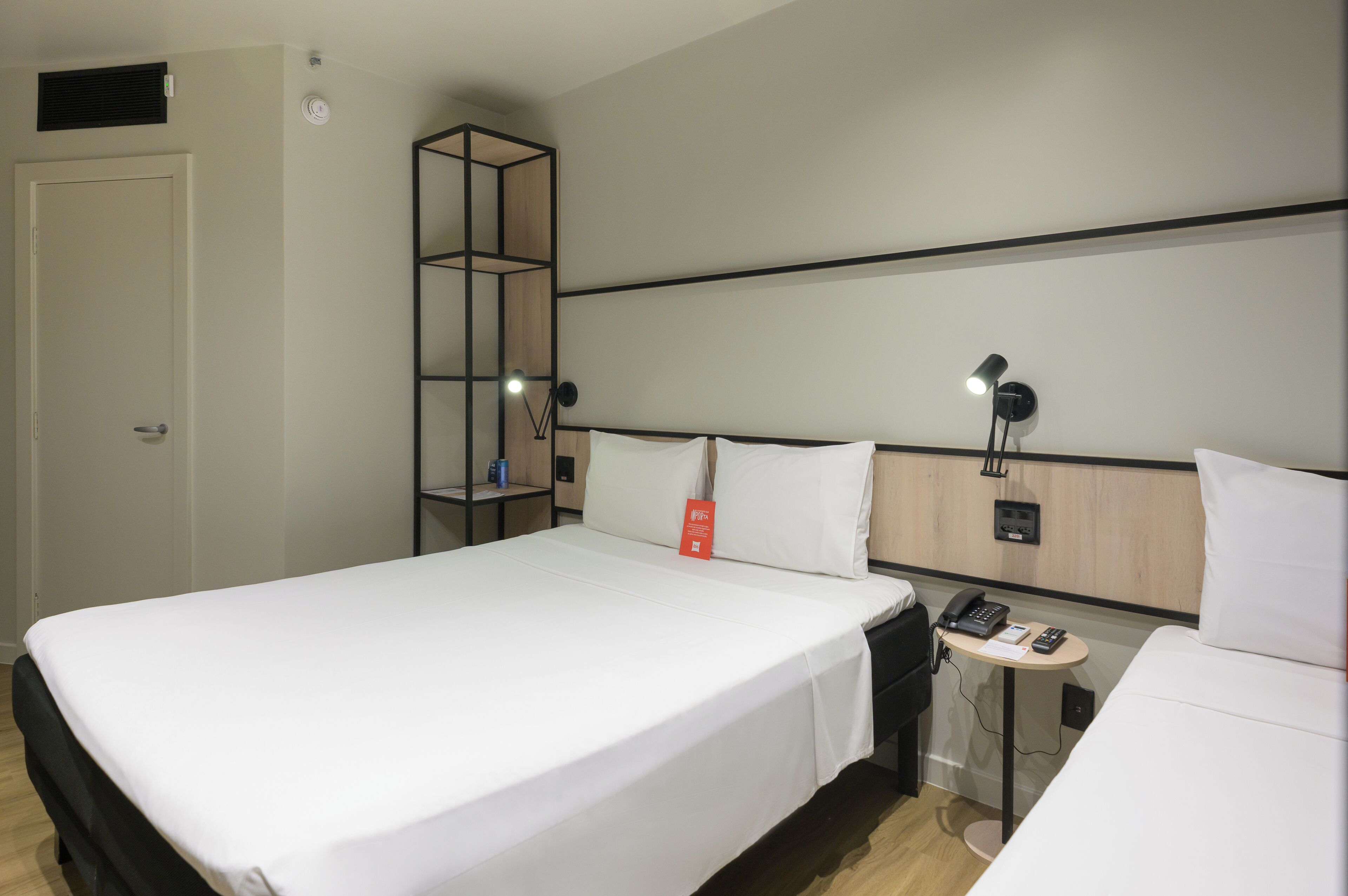 Hotellihuone, jossa on suuri sänky, yöpöytä lampun kanssa, puhelin ja pieni hylly, jossa on pullo.