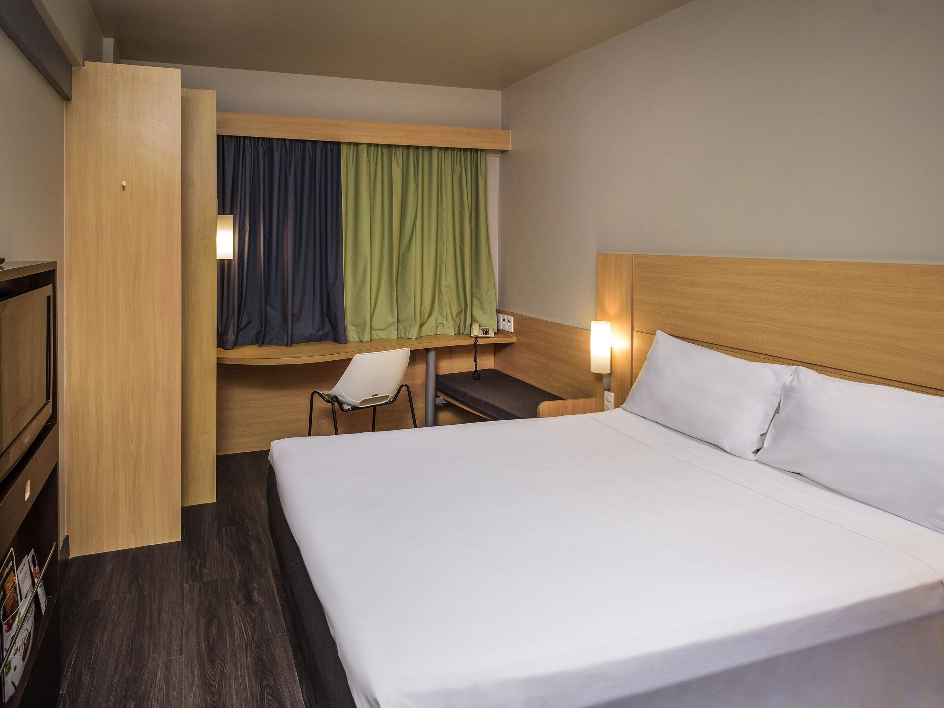 A hotel room with a bed, a desk, a chair, a TV, and a window with curtains.
