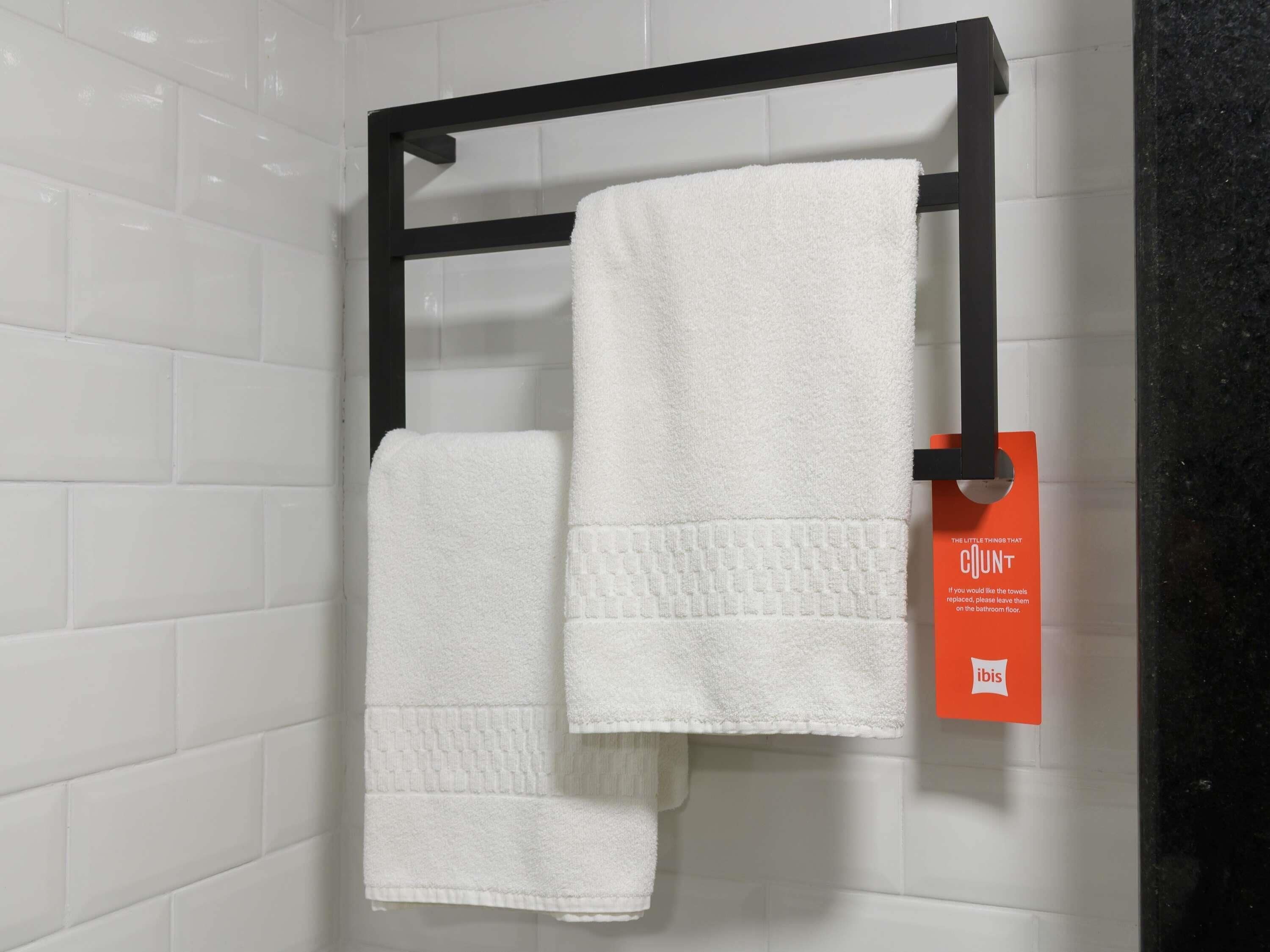 Un baño con paredes de azulejos blancos, un toallero negro que sostiene dos toallas blancas y una etiqueta naranja con texto.