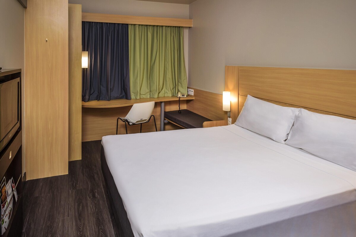 Ein Hotelzimmer mit Bett, Schreibtisch, Stuhl, Fernseher und einem Fenster mit Vorhängen.