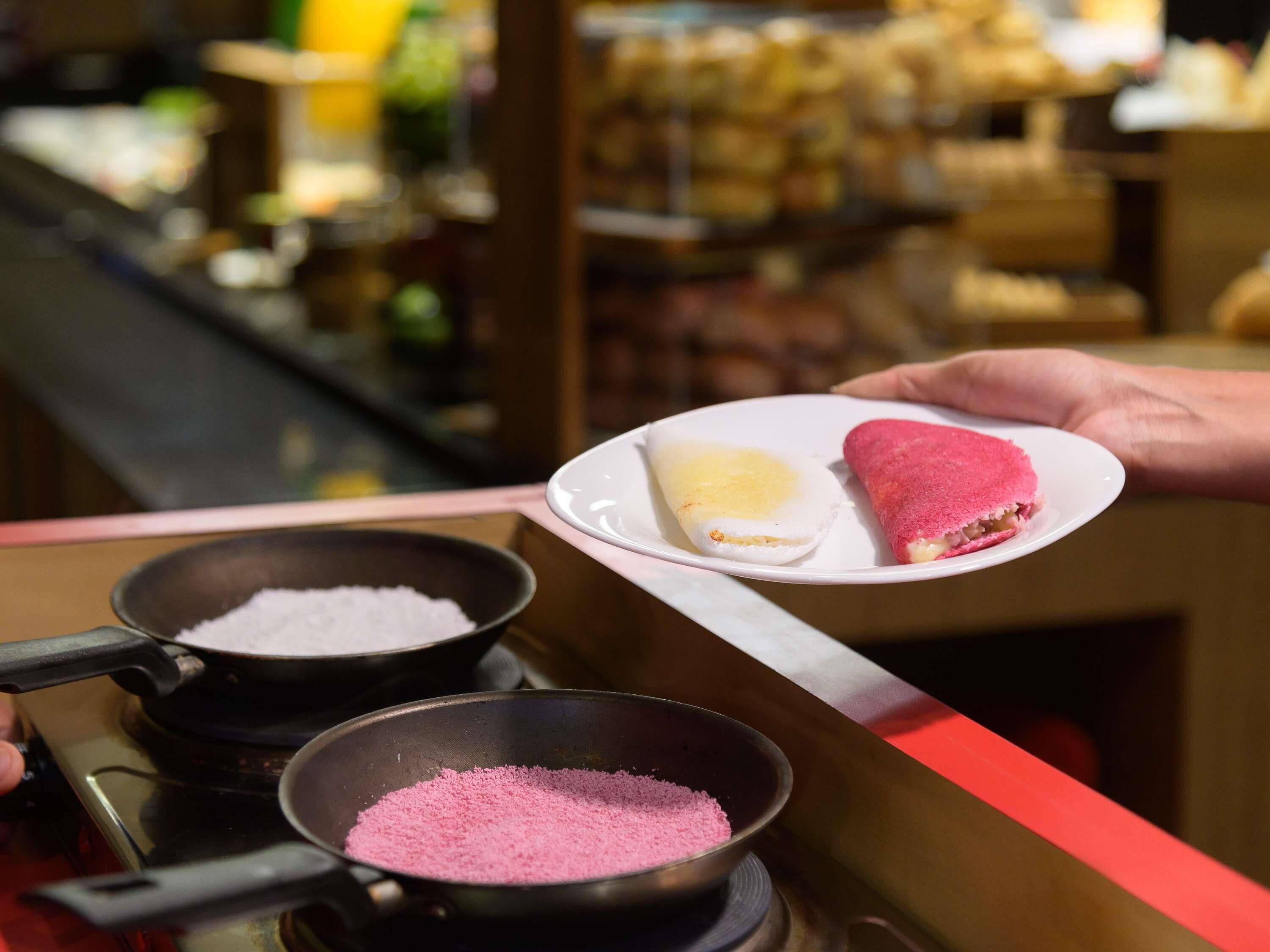 A person holding a plate with two food items, one with a yellow filling and another with a pink filling, over a counter with two pans, one containing pink powder.