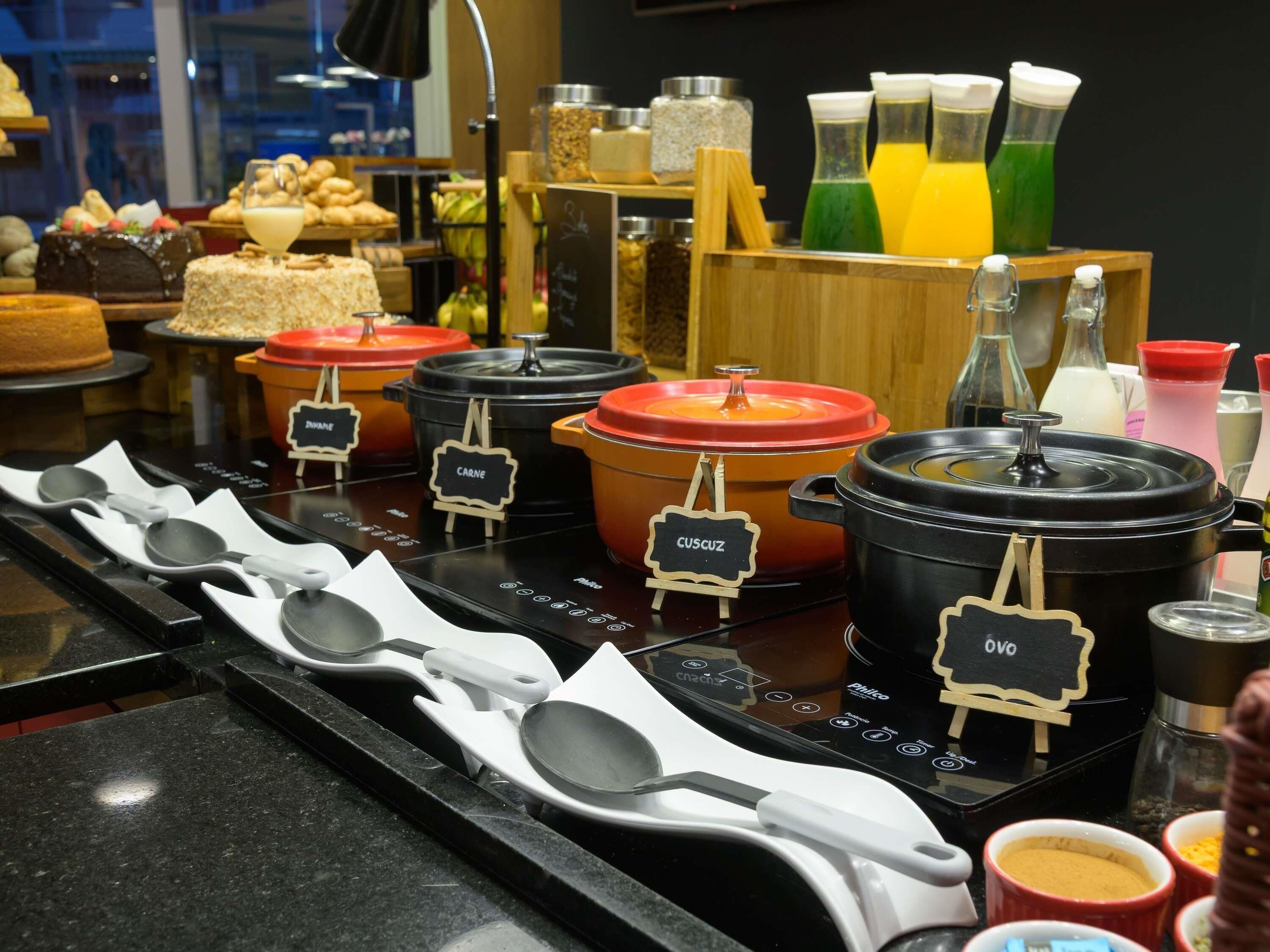 Uma área de buffet com diversos recipientes de comida identificados com diferentes pratos como 'Carne', 'Cucuz' e 'Ovo'.