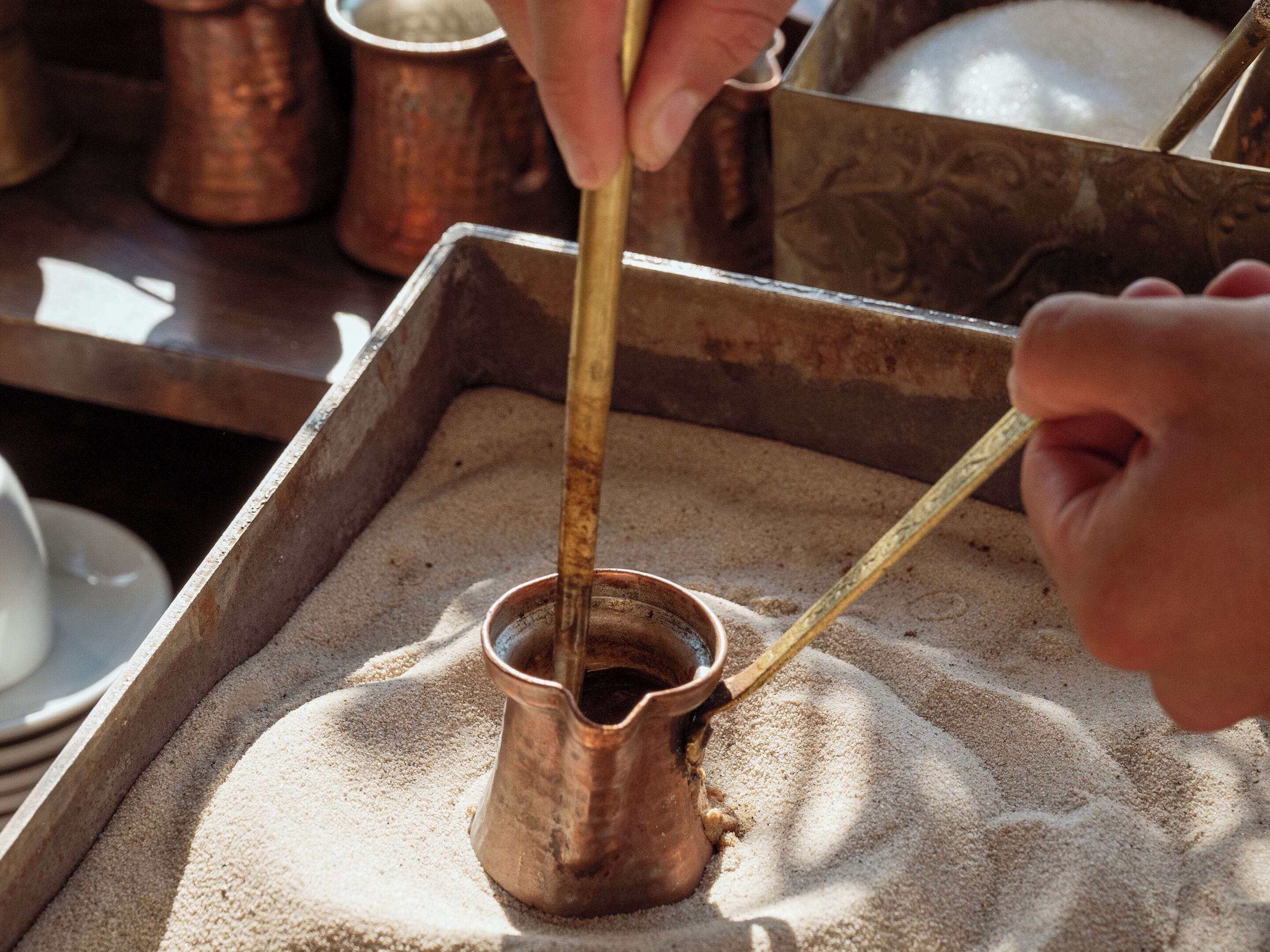 Una olla de cobre sobre una bandeja con arena, una mano sosteniendo un palo, y un fondo borroso con un recipiente de cobre.