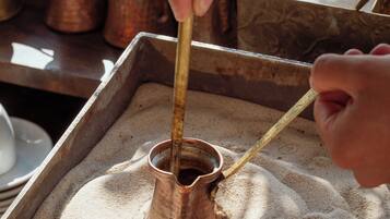 Una olla de cobre sobre una bandeja con arena, una mano sosteniendo un palo, y un fondo borroso con un recipiente de cobre.