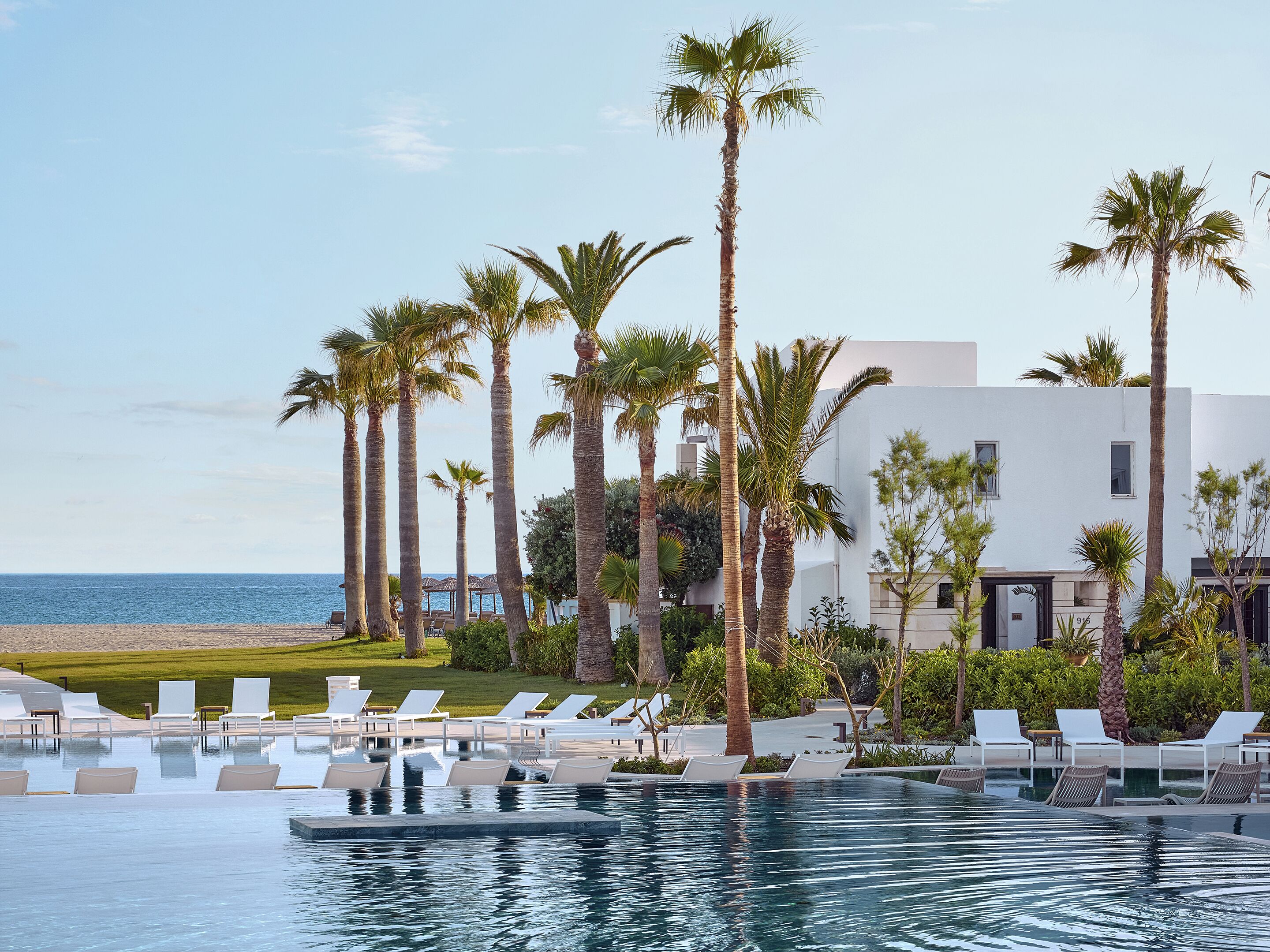 Ein Poolbereich mit Liegestühlen und Palmen, mit Blick auf einen Strand und ein modernes Gebäude.