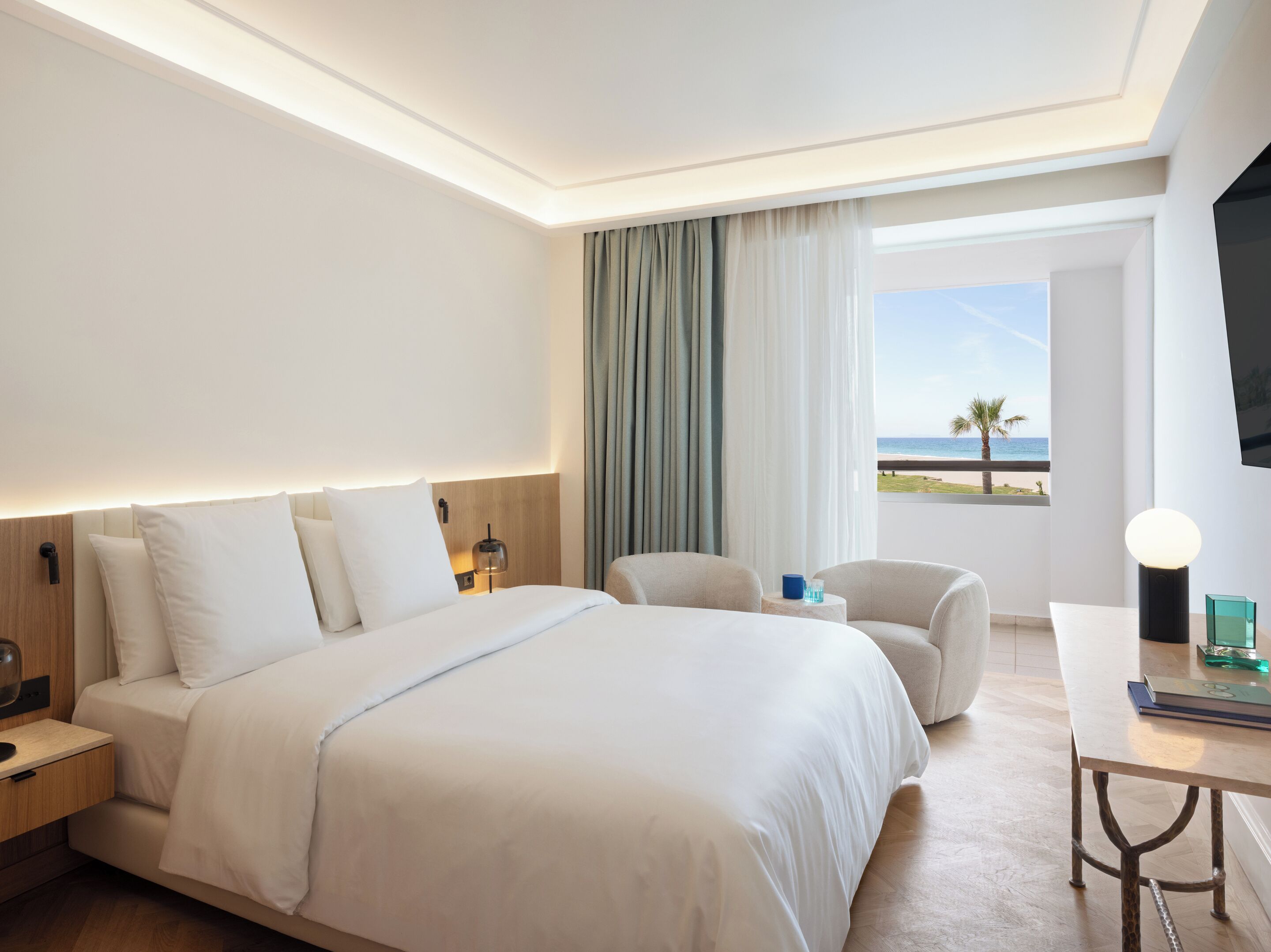 A hotel room with a large bed, a desk, a chair, a TV, and a view of the beach.