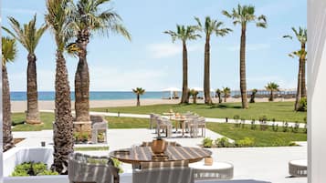 A terrace with outdoor seating, palm trees, and a view of the beach.