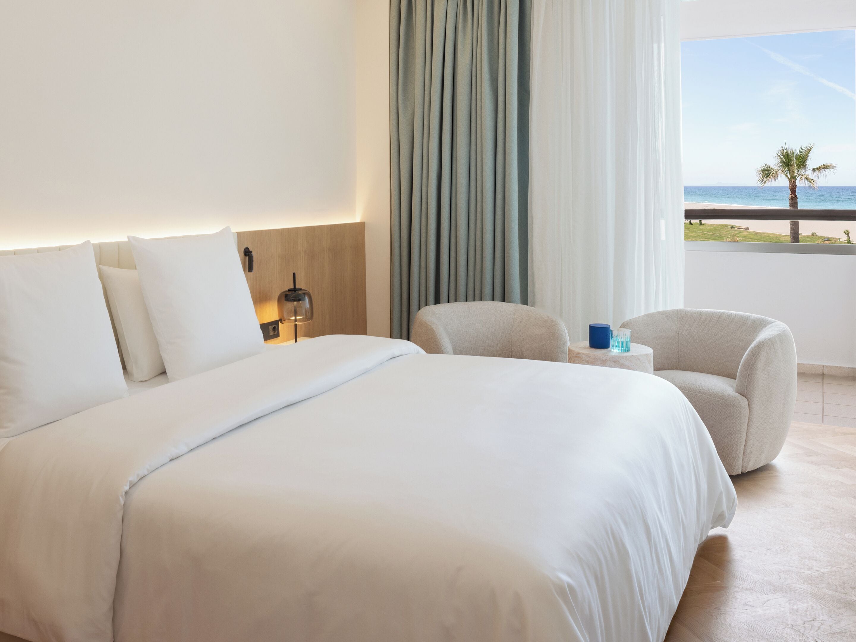 A hotel room with a large bed, a seating area with a chair, a nightstand, and a view of the ocean.