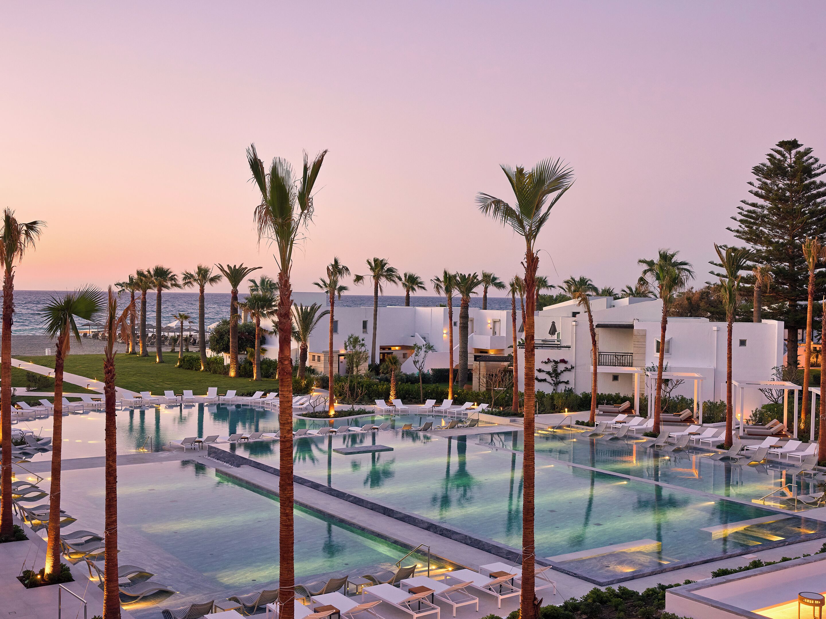 Een resort met een groot zwembad, palmbomen en uitzicht op zee bij zonsondergang.