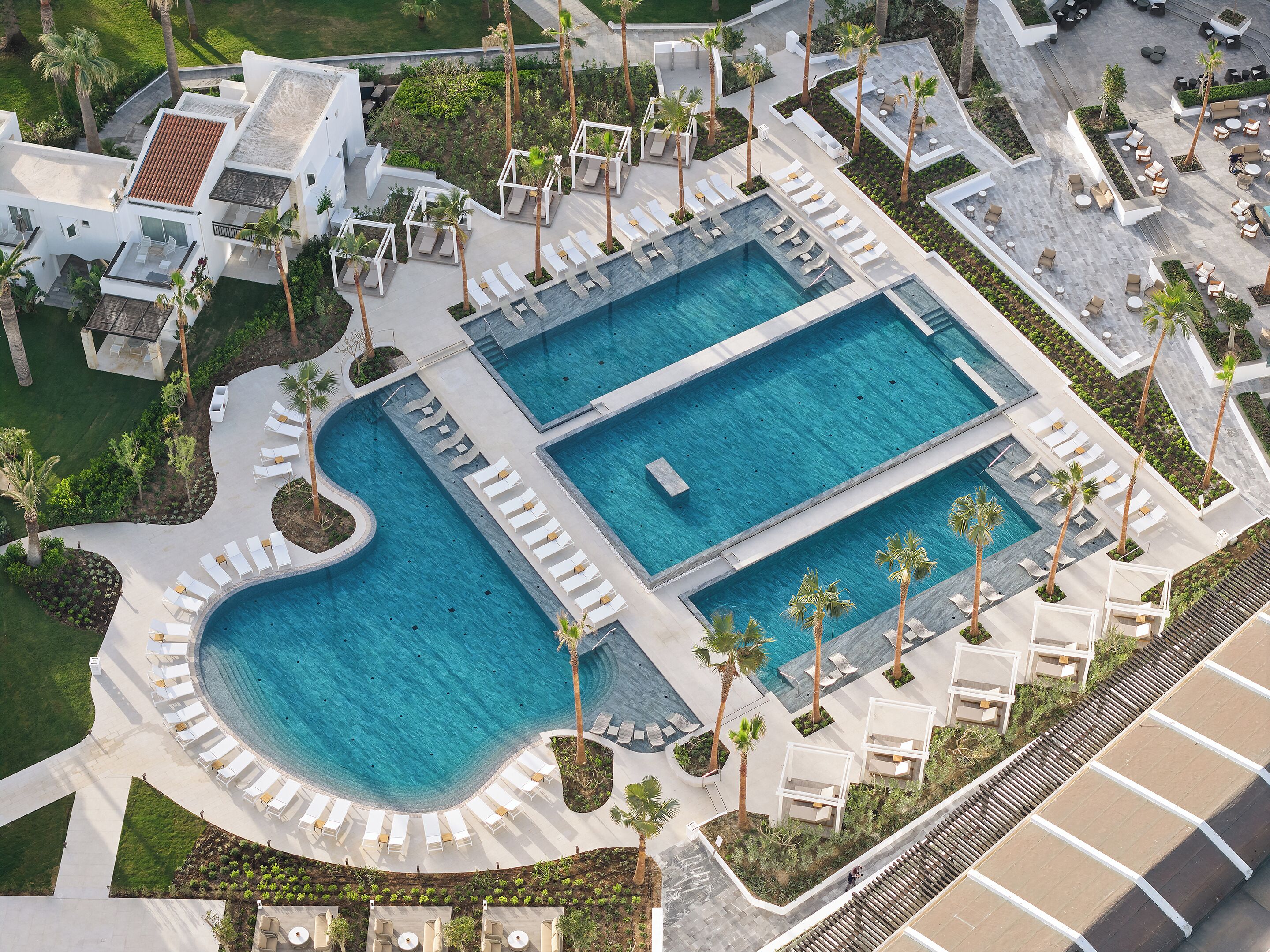Vista aérea de un complejo turístico con dos piscinas grandes, tumbonas, palmeras y un diseño arquitectónico moderno.