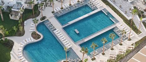 Vista aérea de un complejo turístico con dos piscinas grandes, tumbonas, palmeras y un diseño arquitectónico moderno.