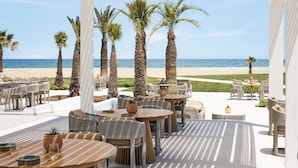 Un espacio al aire libre techado con mesas y sillas, palmeras y vista a la playa.