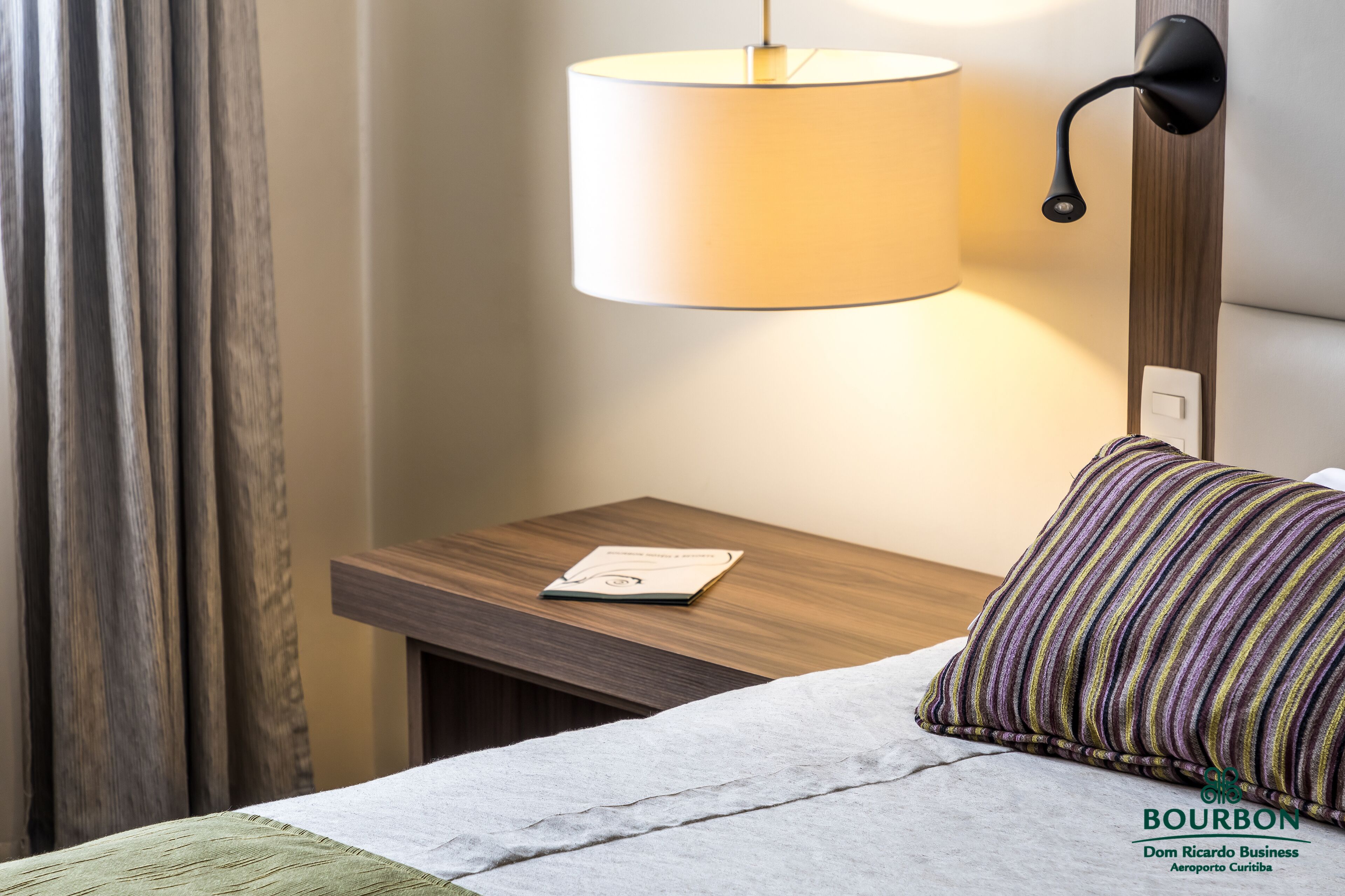 A hotel room with a bed, a nightstand with a lamp, a pillow, and a wall-mounted switch.