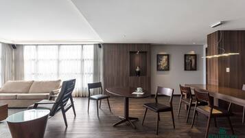 Una sala moderna con un sofá, una mesa de comedor redonda y sillas. Hay ventanas amplias con cortinas, piso de madera y una lámpara de techo fijada a la pared.