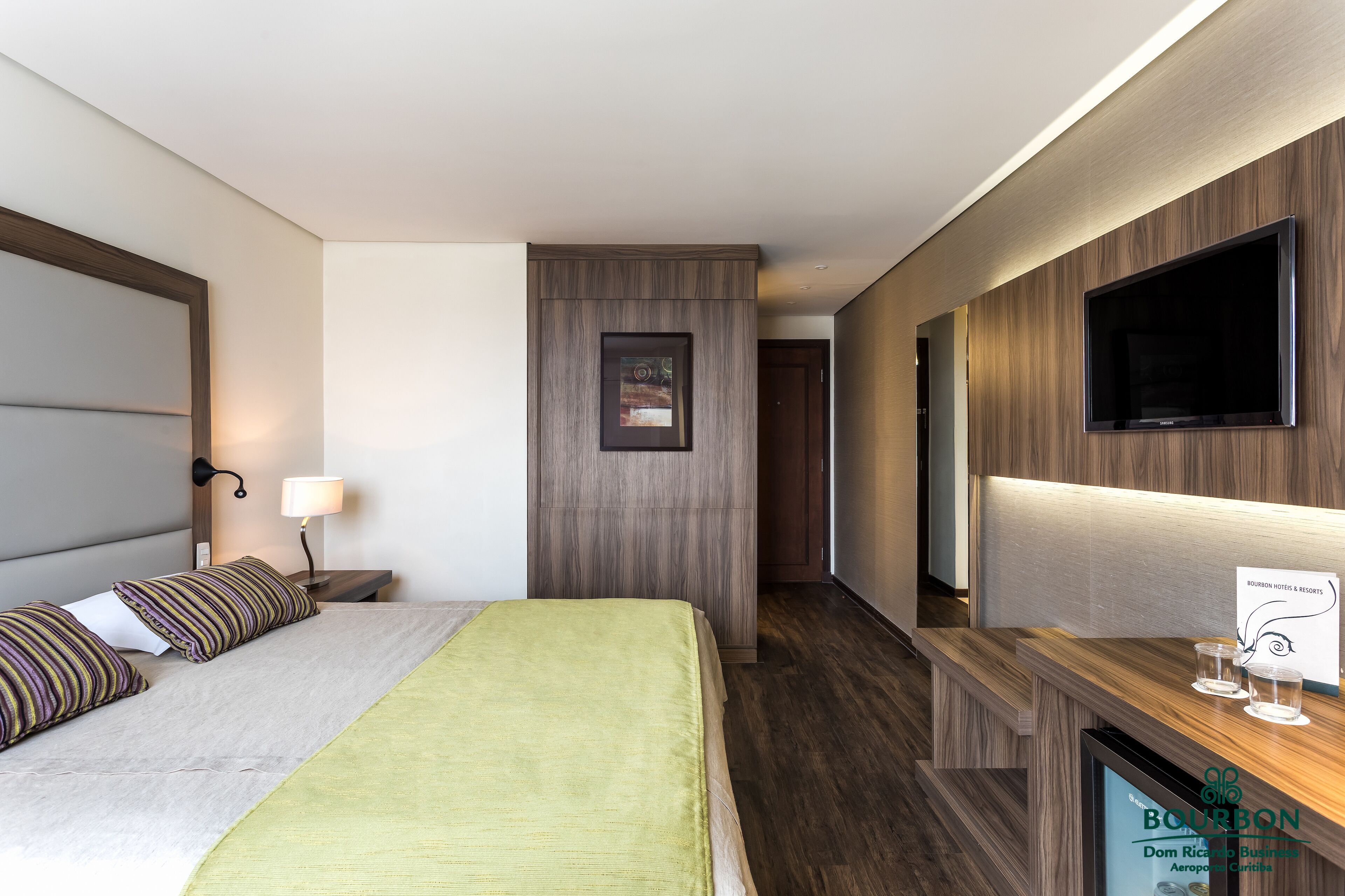 A modern hotel room with a bed, bedside lamp, and a flat-screen TV on a wooden panel wall.