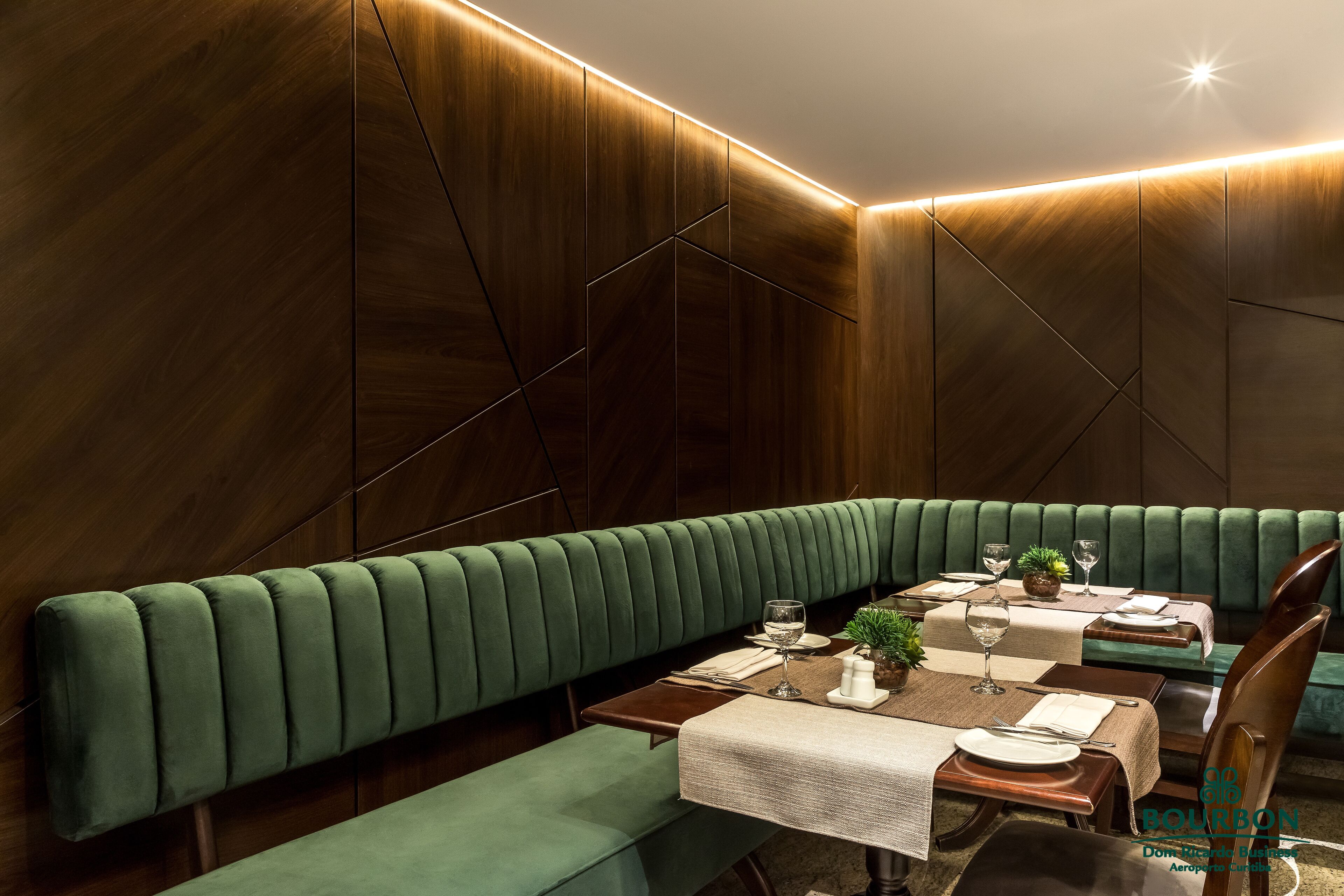 A modern dining area with a long green bench, wooden panel walls, and a table set with plates, glasses, and a small plant.