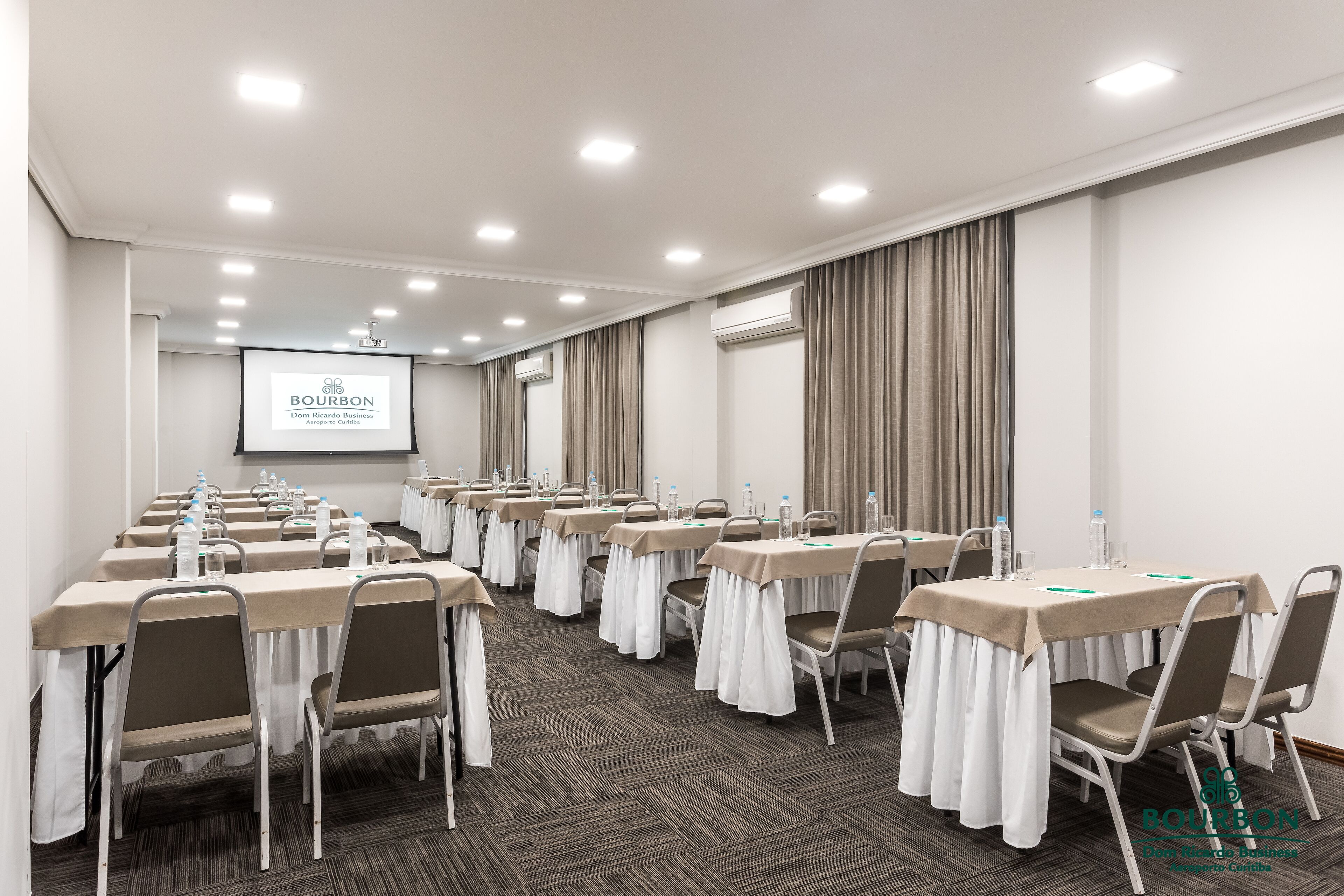 Sala de conferências com várias mesas, cadeiras e uma tela de projeção.
