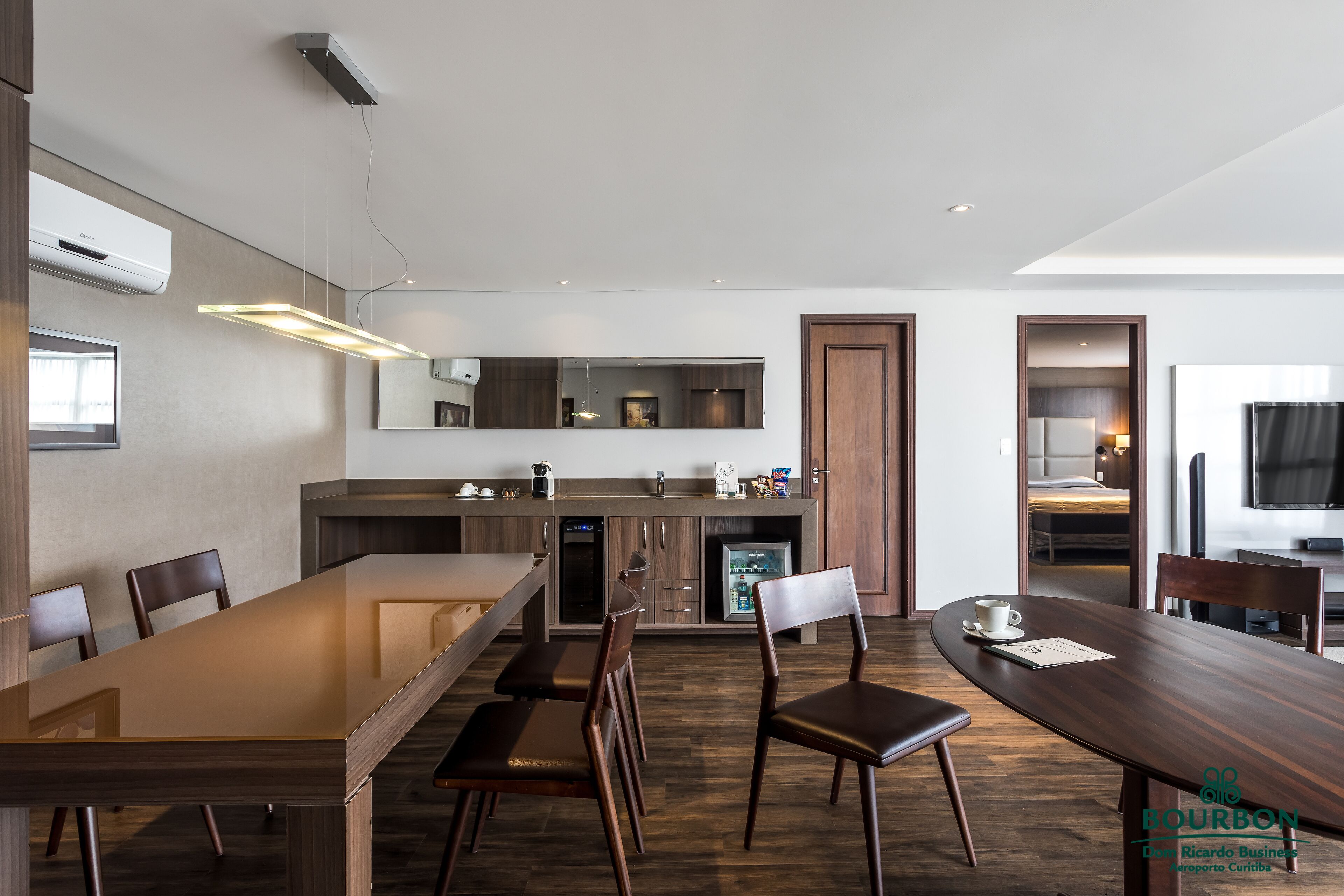 Habitación de hotel moderna con una gran mesa de madera, varias sillas, una cocina integrada y un espejo grande.