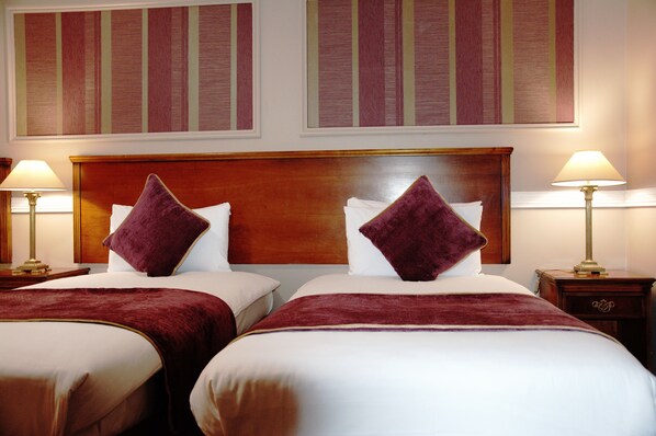 A hotel room with two beds, each with a wooden headboard, white linens, and a red throw blanket. There are two bedside tables with lamps, and the walls have striped wallpaper.