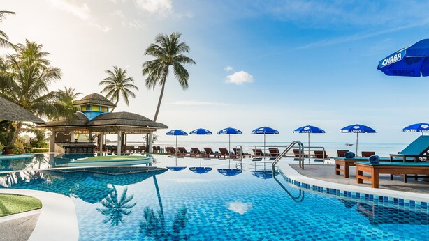A swimming pool with lounge chairs and umbrellas, surrounded by palm trees and a beach view.