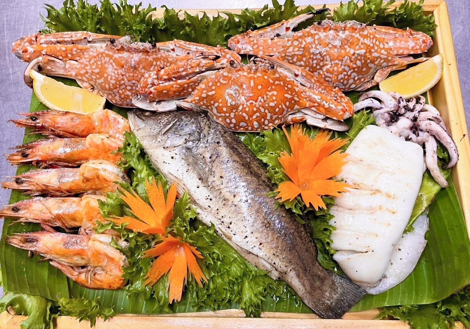 A seafood platter with crabs, shrimp, fish, and squid, garnished with lemon and carrot slices on a banana leaf.