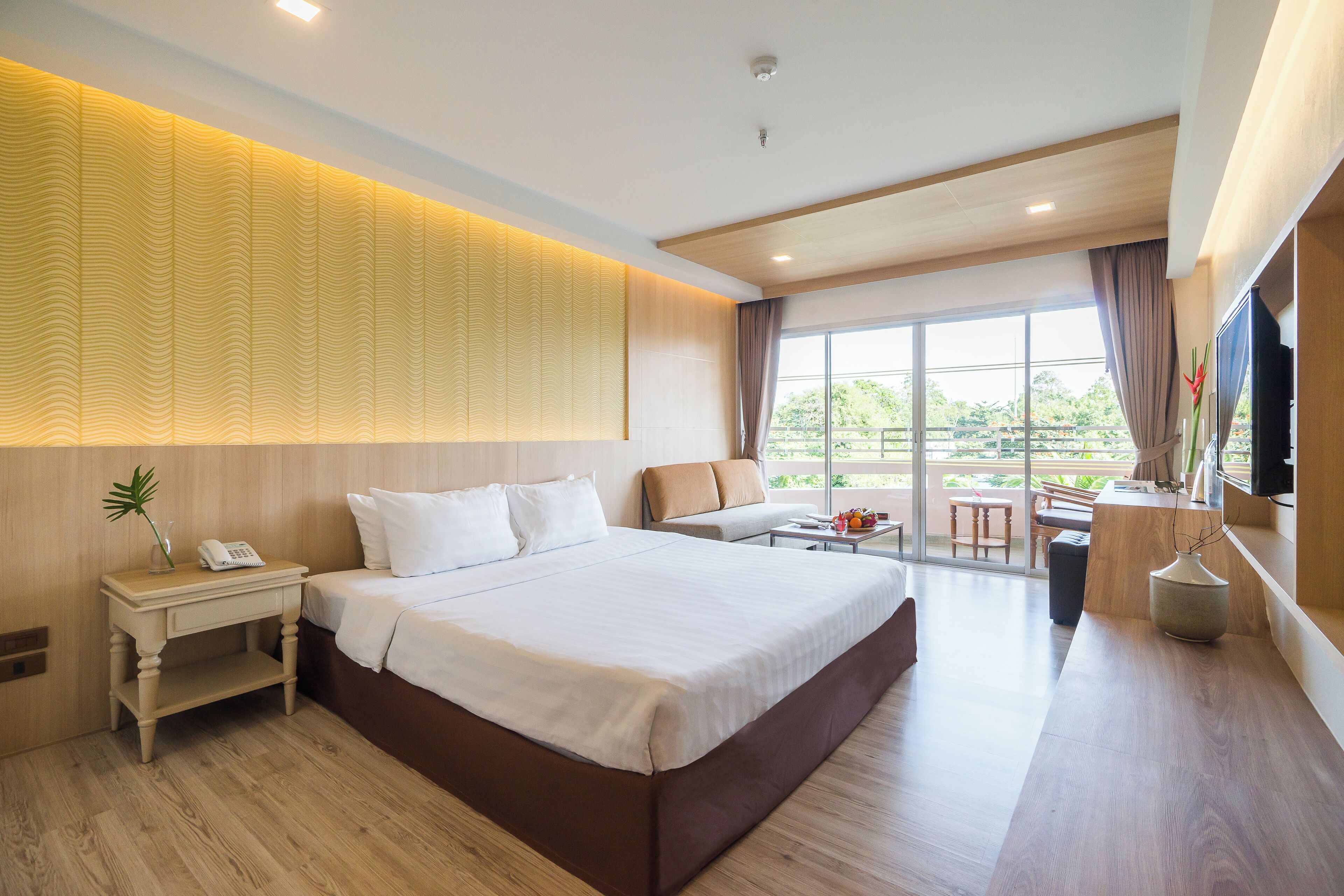 A modern hotel room with a large bed, a bedside table with a phone, a sofa, a small table with a vase, and a view of the outdoors through large windows.