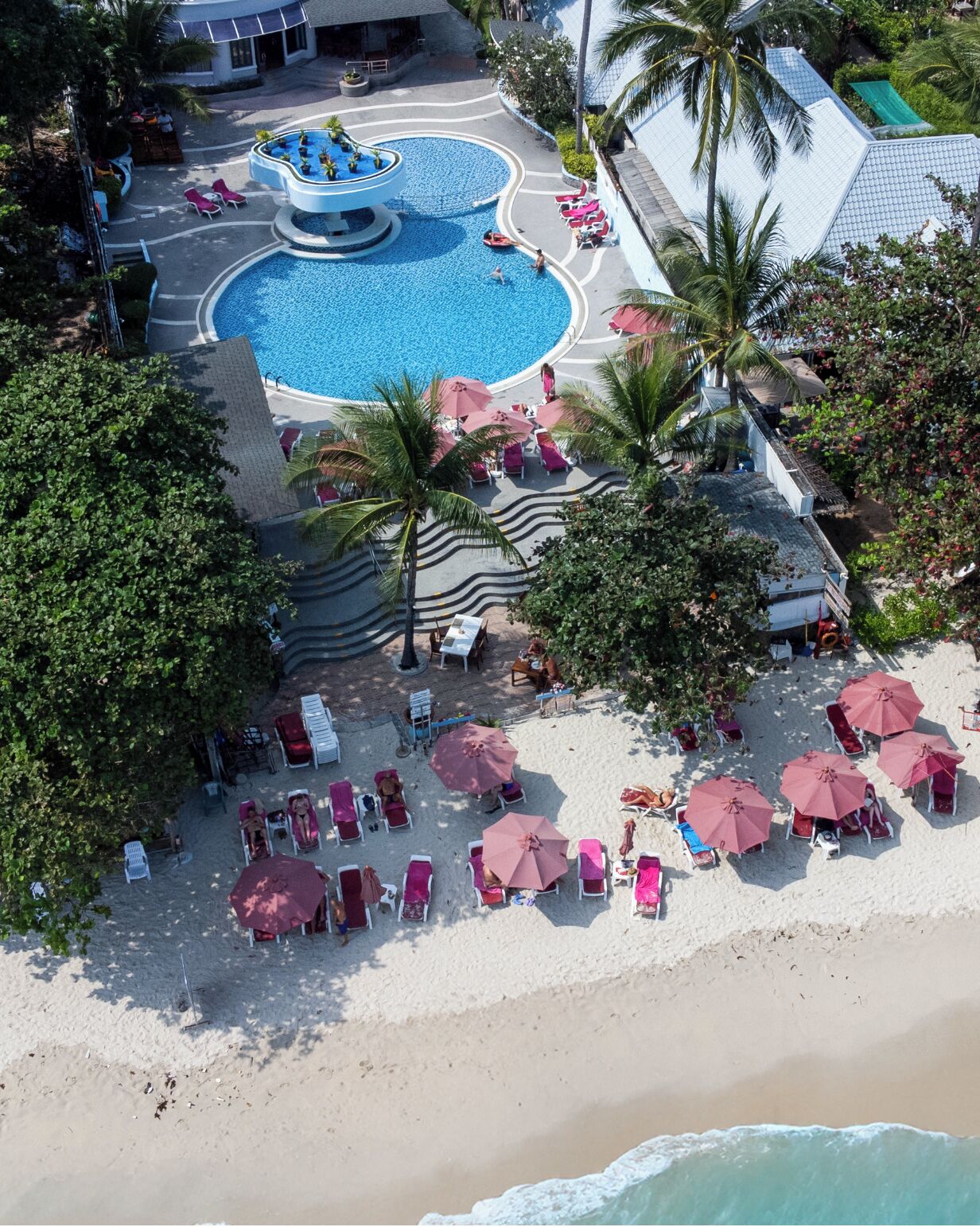 An aerial view of a resort with a central swimming pool, multiple beach areas with sun loungers and umbrellas, and palm trees.