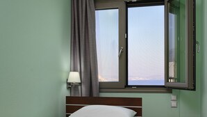 A hotel room with a bed, a nightstand, a lamp, a window with curtains, and a view of the sea. - Avra (Agios Vasileios)