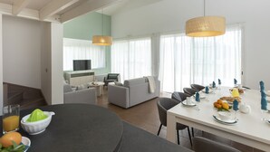 Un salon moderne avec un coin repas, une cuisine à l’arrière-plan et de grandes fenêtres ornées de rideaux légers.