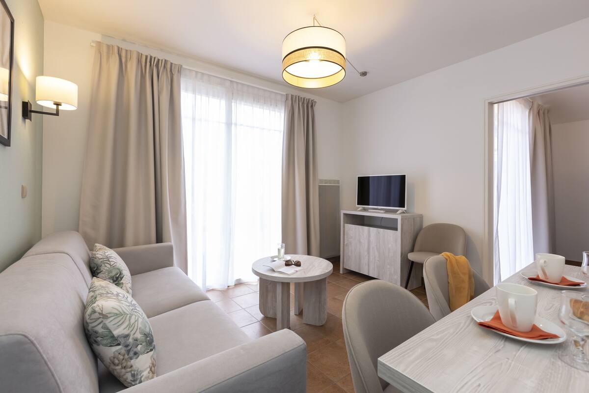 Ein modernes Wohnzimmer mit einer Couch, einem Esstisch mit Tassen und einem Buch, einem Fernseher und einem kleinen Beistelltisch.