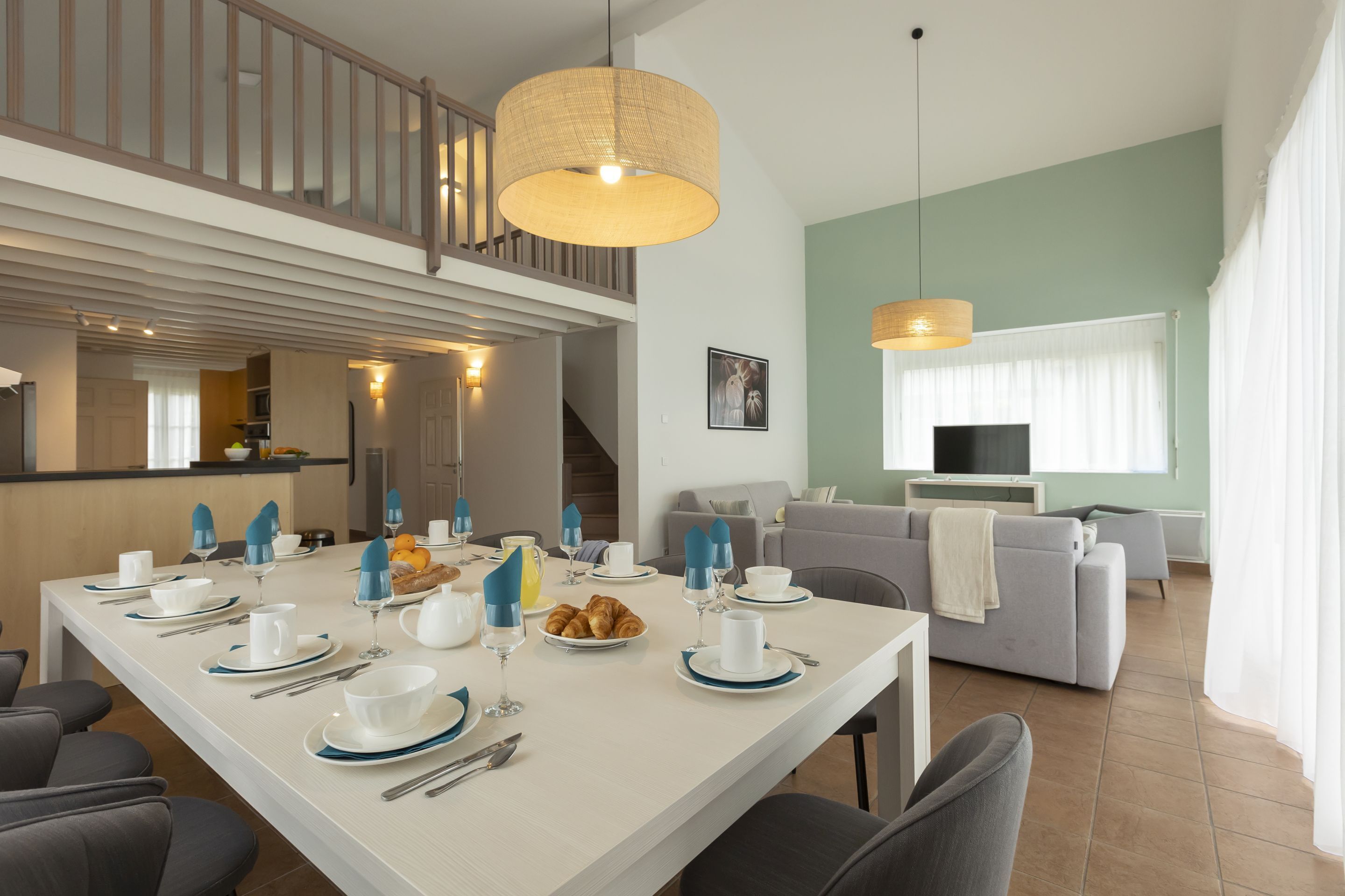 A modern dining area with a long table set for breakfast, a staircase, and a living area with a sofa and TV.