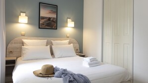 Un lit bien fait, avec du linge de lit blanc, un chapeau de paille et une serviette pliée posée sur le côté. Il y a deux lampes de chevet et un tableau encadré accroché au mur au-dessus du lit.